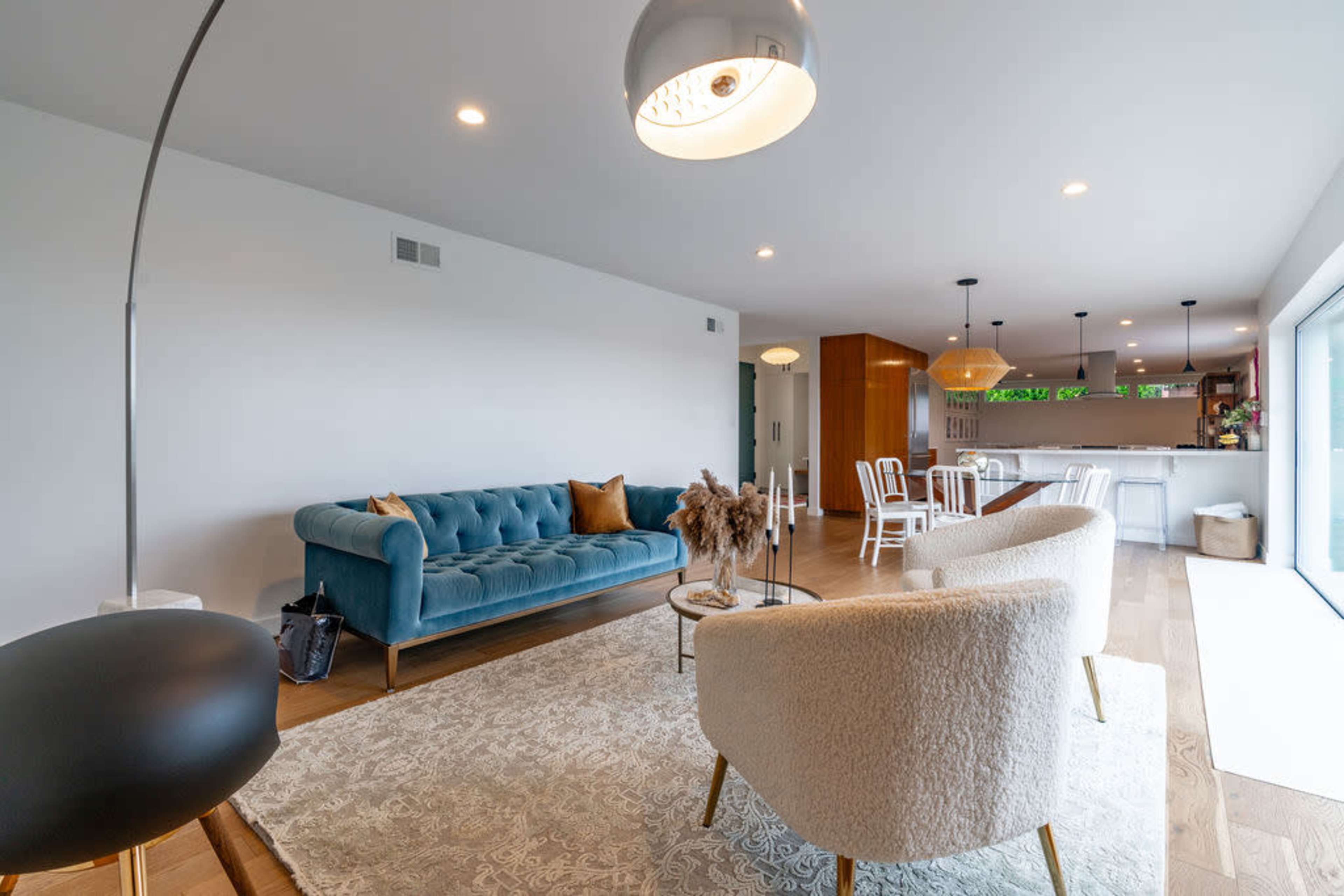 A spacious living area features a blue velvet sofa, two white accent chairs, a coffee table, and an open kitchen in the background.