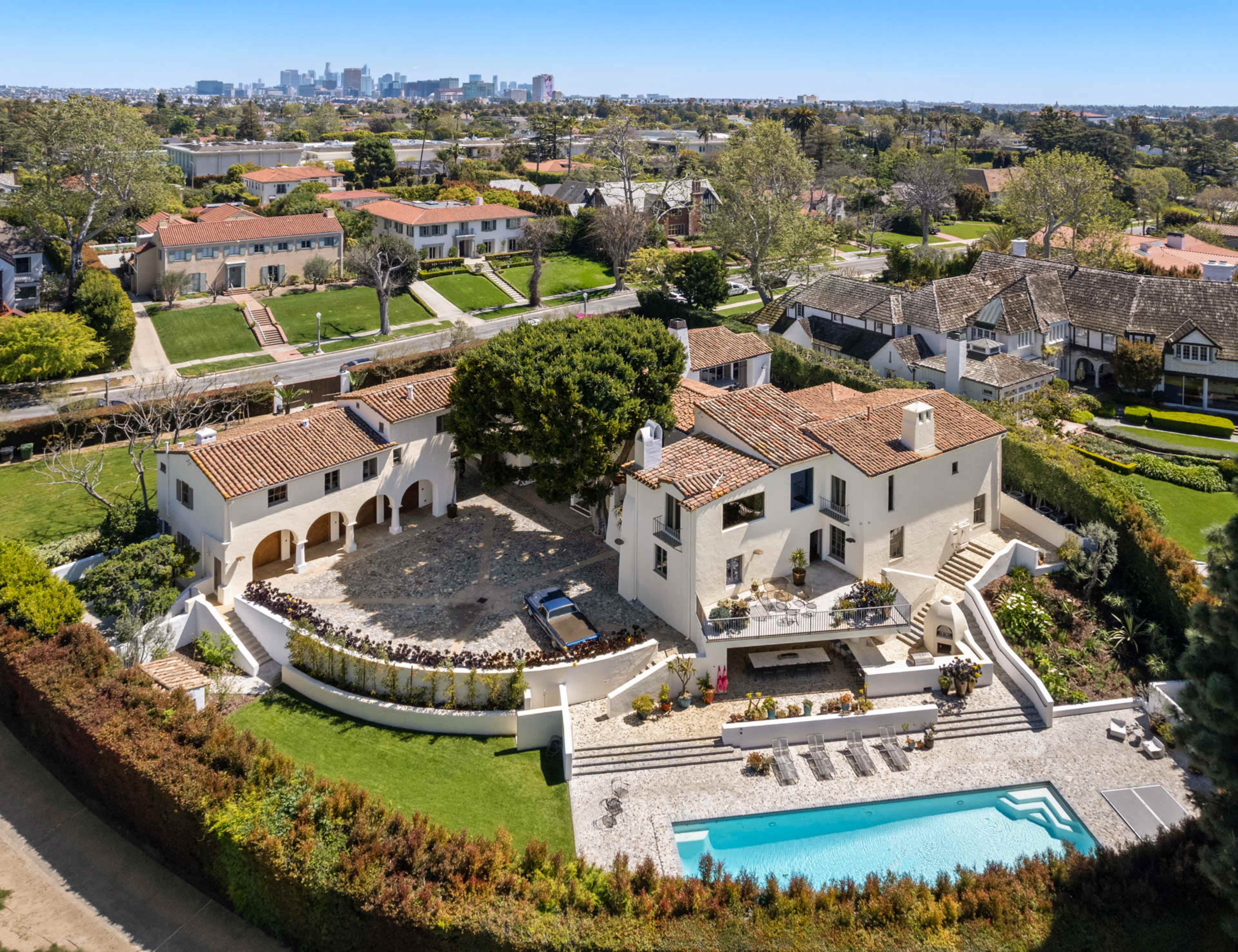 The image shows a large house with a pool, surrounded by landscaped gardens and a view of a city skyline in the background.