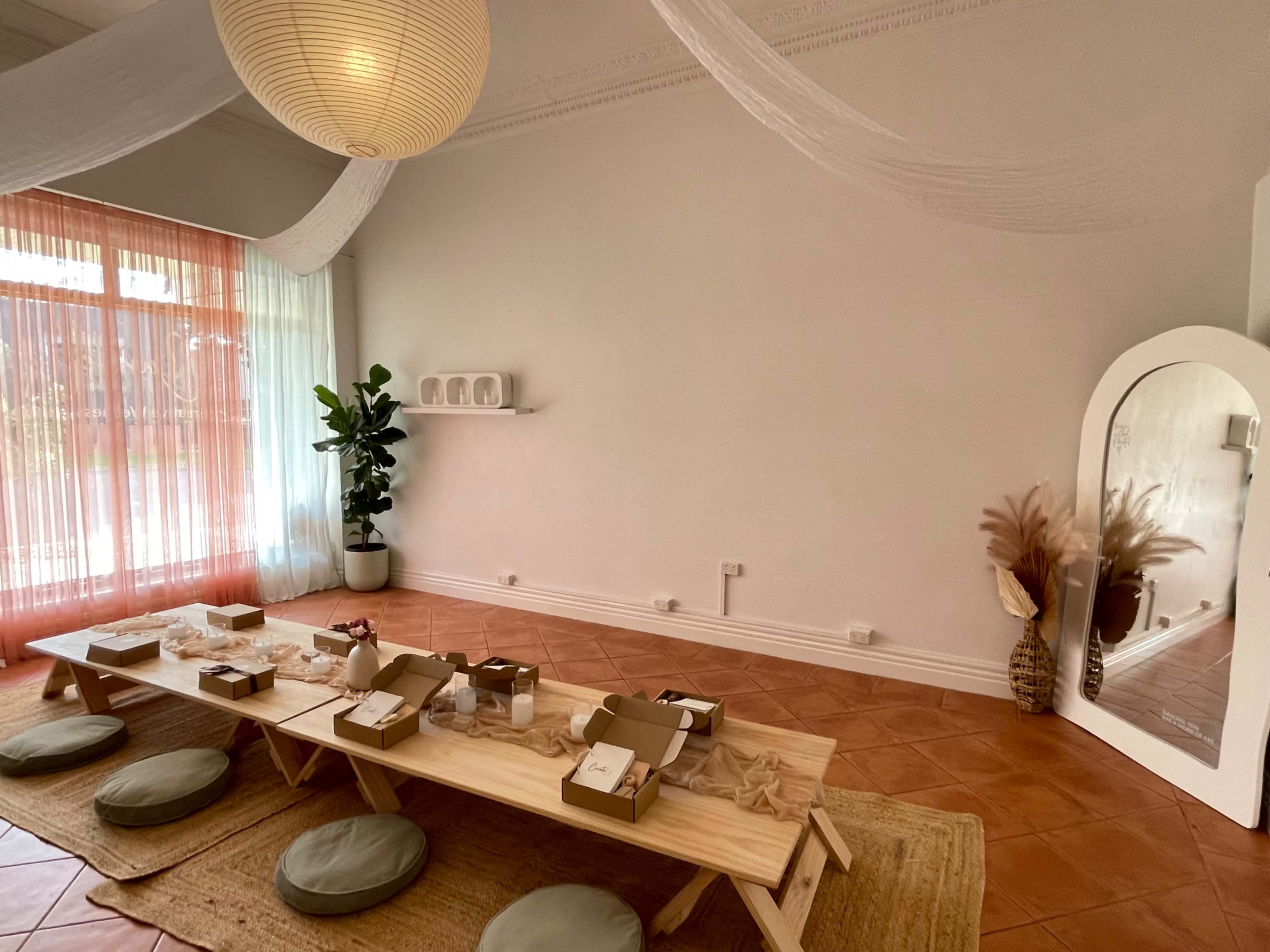 The image shows a spacious, minimalist room with a wooden low table set for a gathering, surrounded by round cushions, plants, and decorative elements.