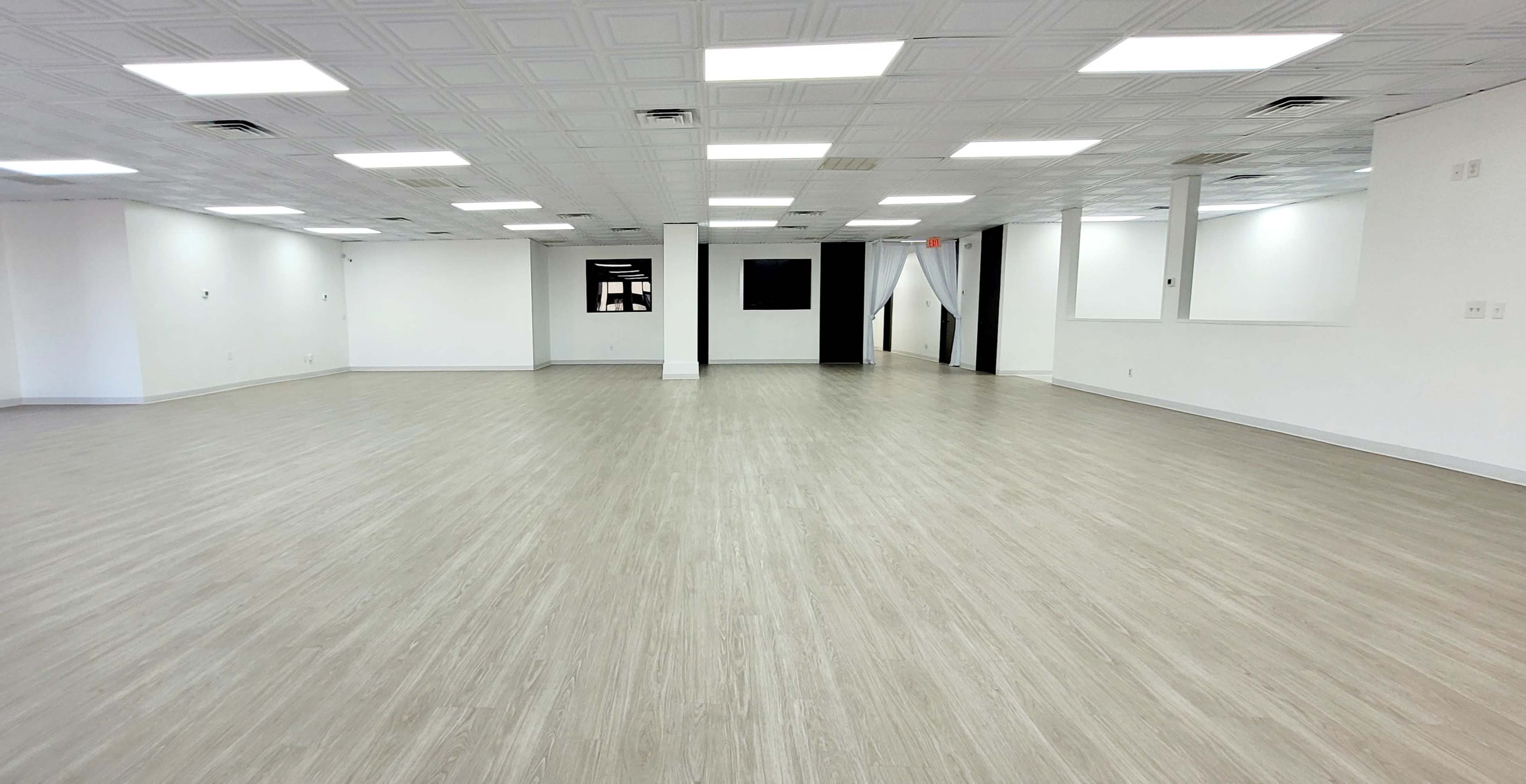 A spacious, empty room with light-colored, wooden flooring and white walls, illuminated by overhead lighting.