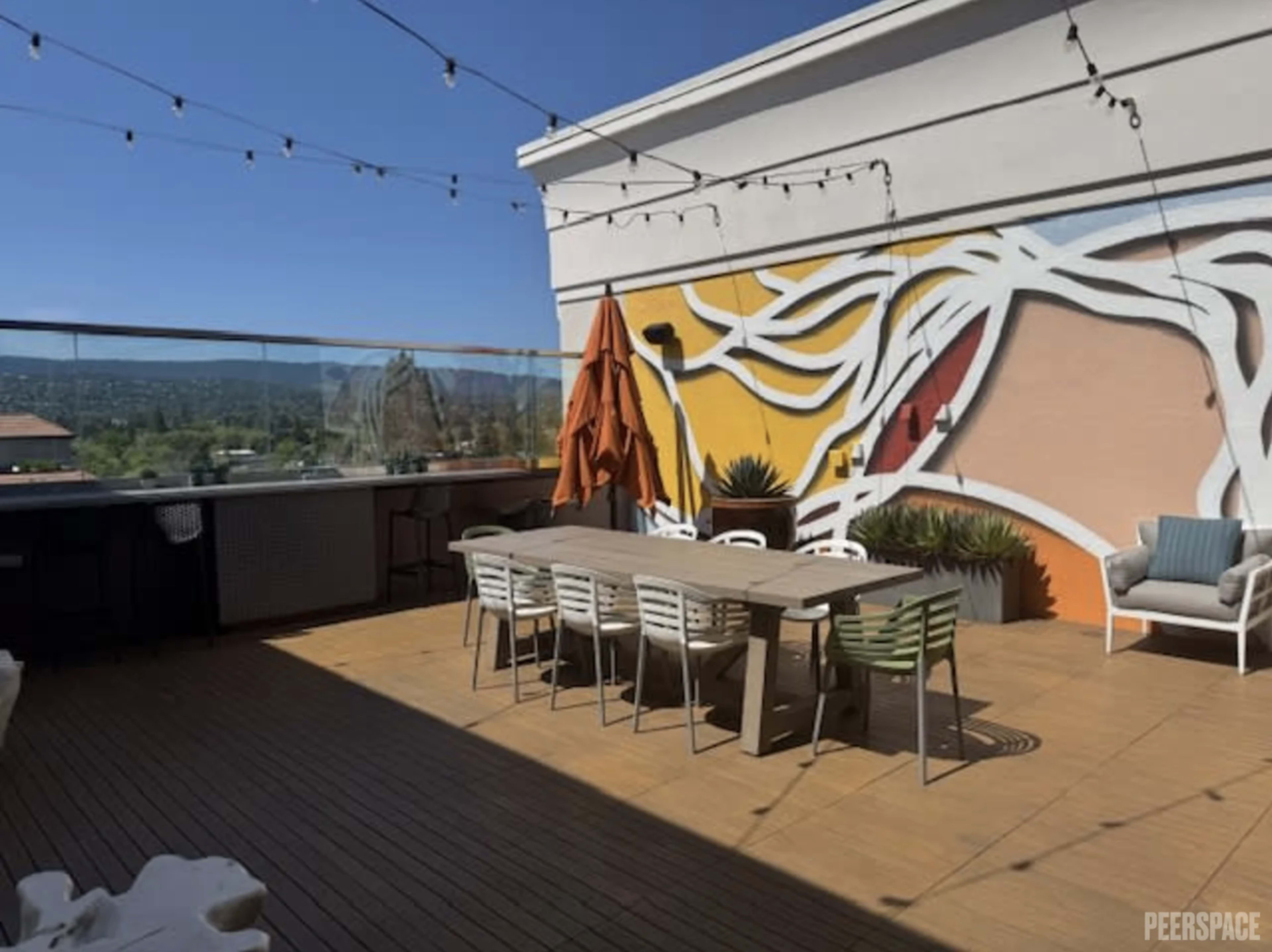 The image shows a spacious rooftop terrace with a large dining table, multiple chairs, a decorative wall mural, and a clear blue sky in the background.