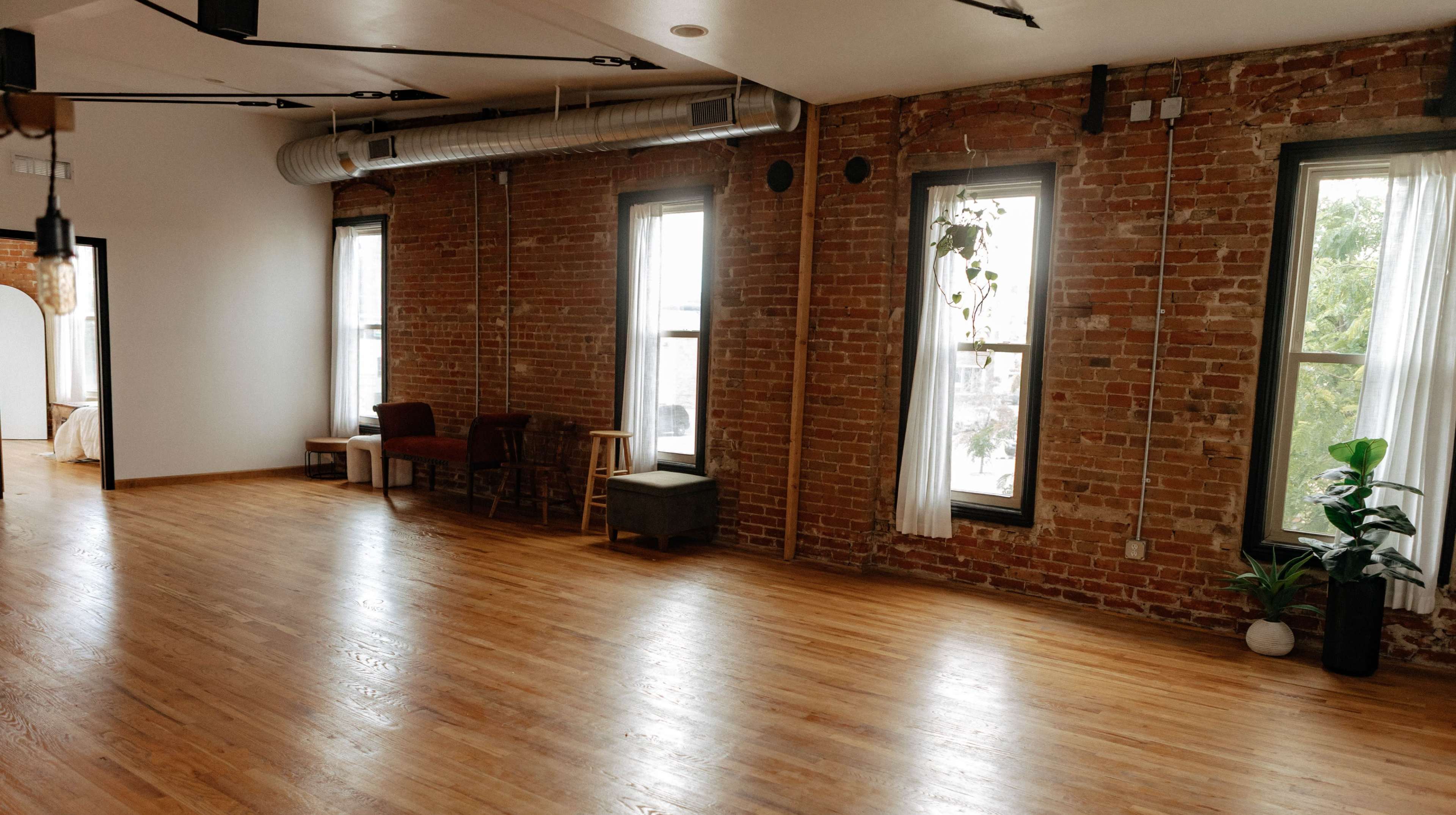 The image shows a spacious room with exposed brick walls, wooden floors, and large windows.