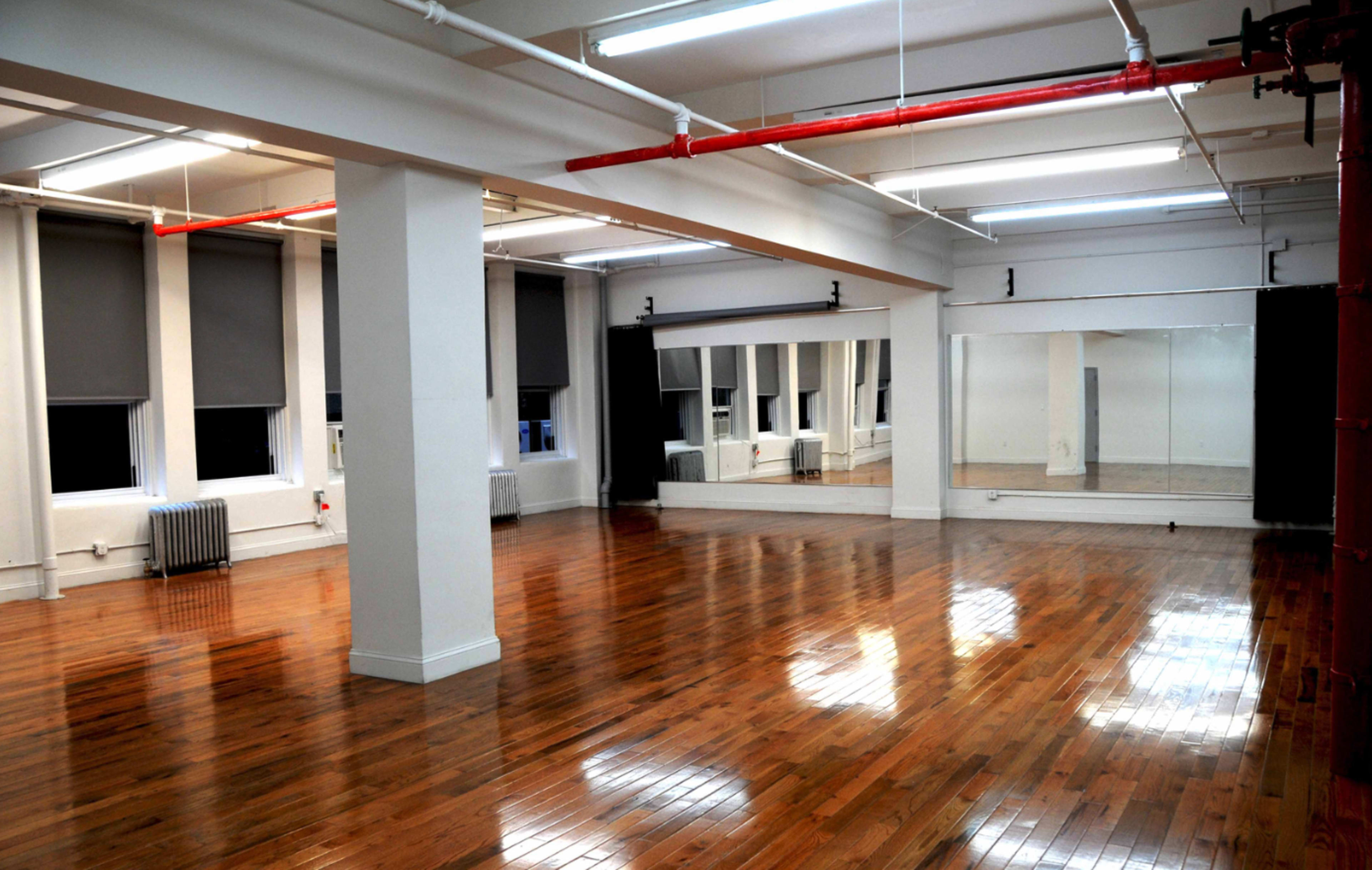 A spacious, empty room with polished wooden floors and large mirrors on one wall, illuminated by overhead fluorescent lights.