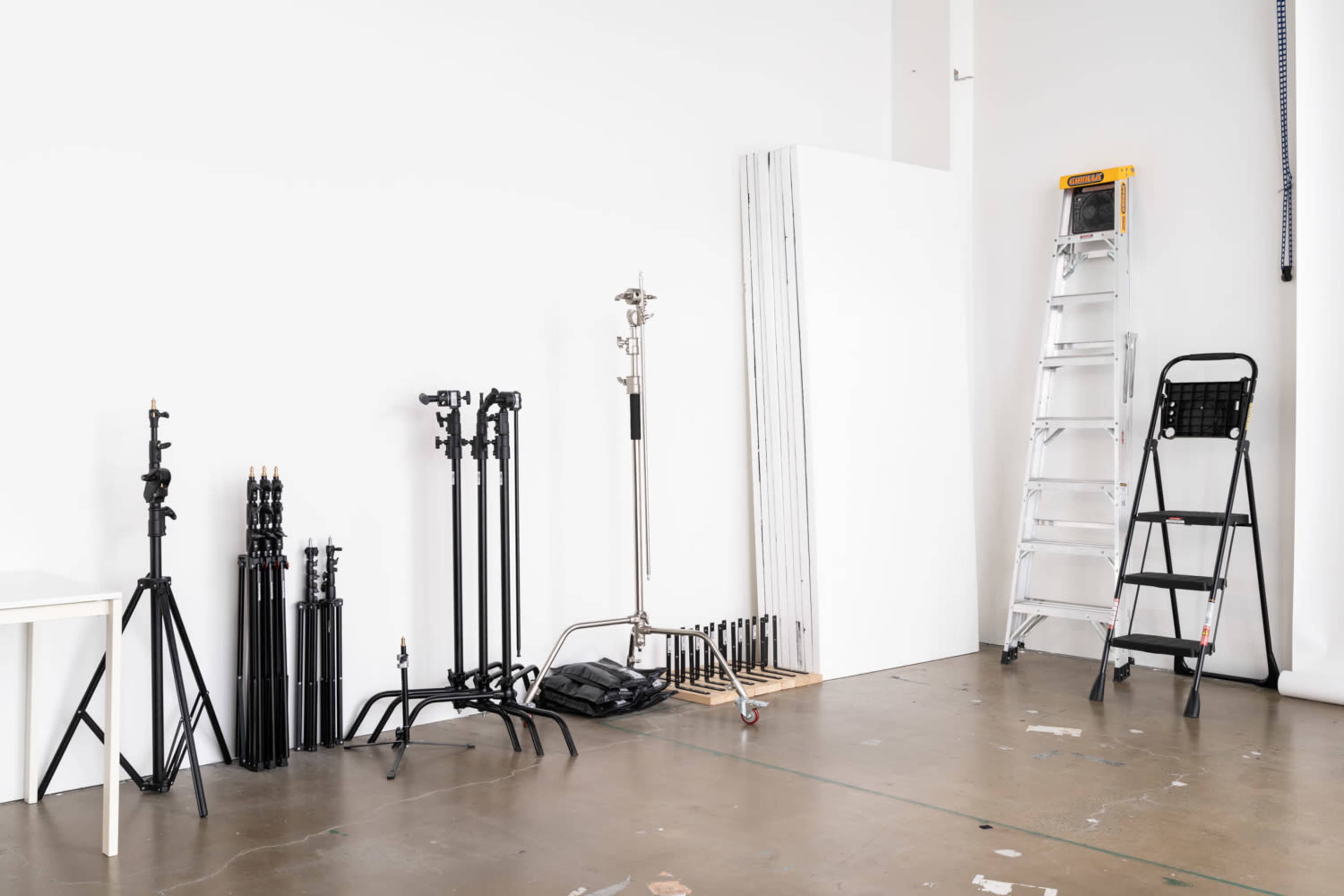 A photography studio with various equipment including light stands, a ladder, and large white panels against a white wall.