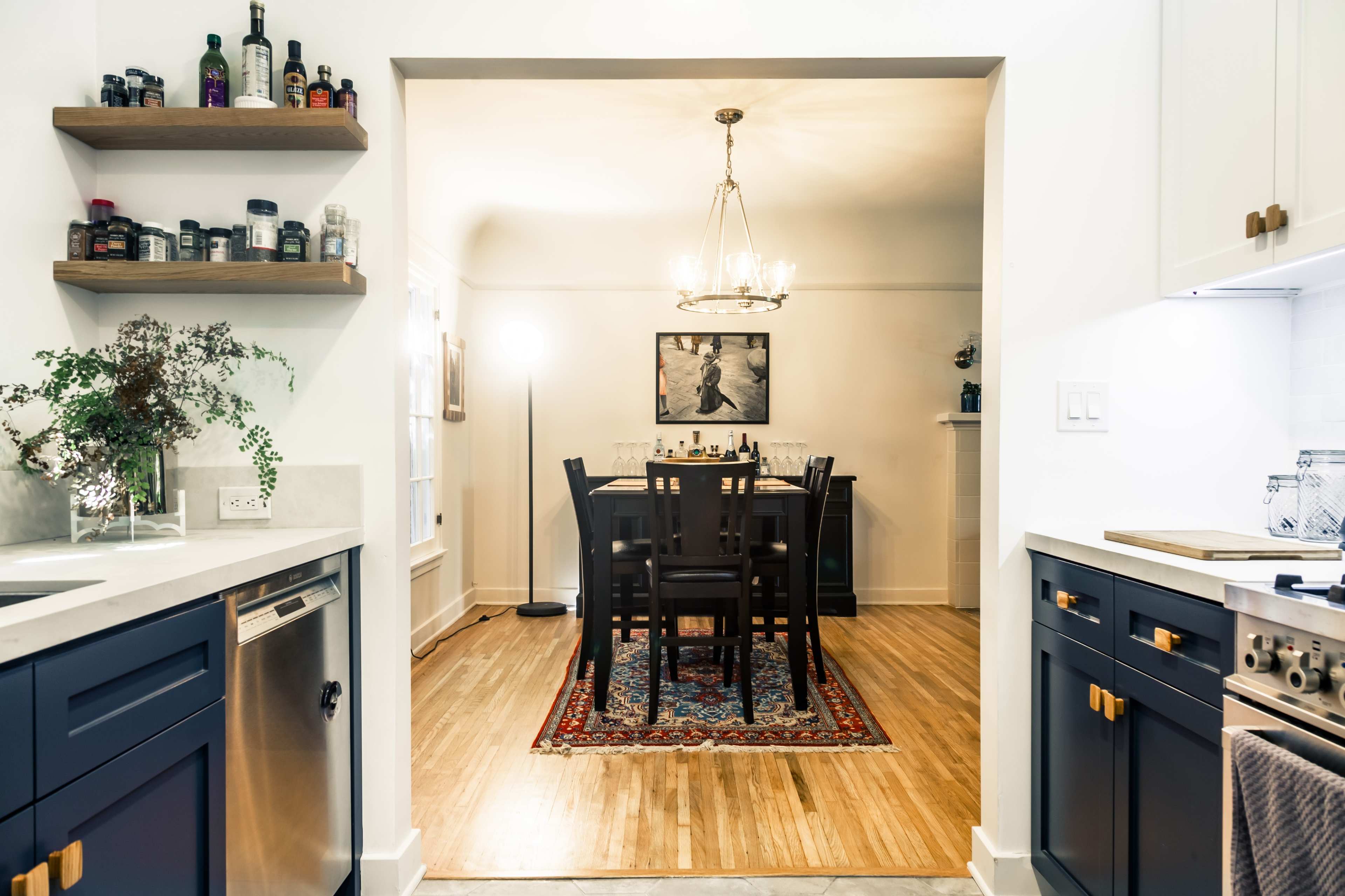 A dining area featuring a table set for four, illuminated by a chandelier, is seen through an open doorway from a kitchen with dark cabinetry and decorative shelves.