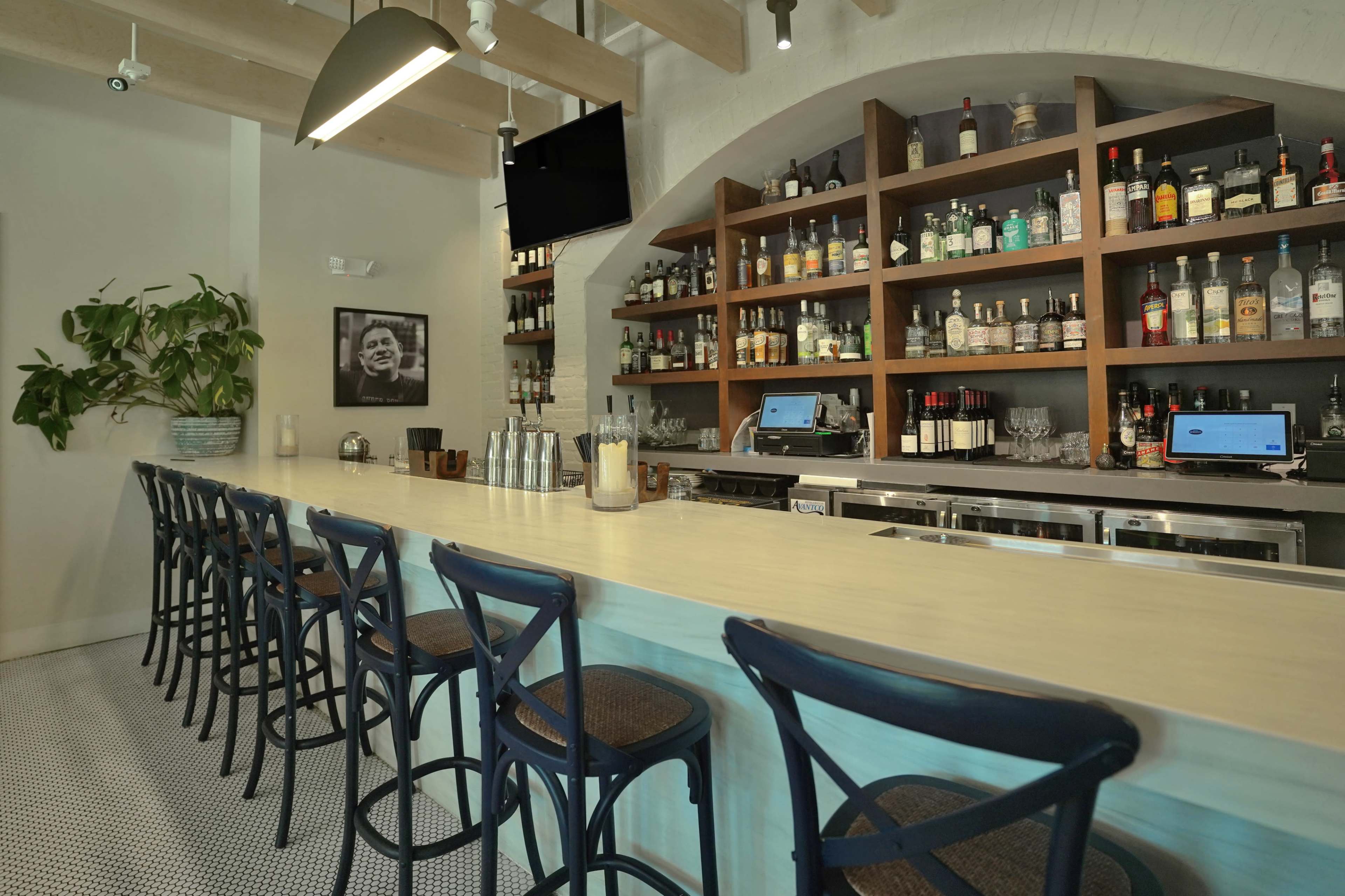 The image shows a modern bar area with a long counter, high stools, and shelves lined with various bottles of liquor.