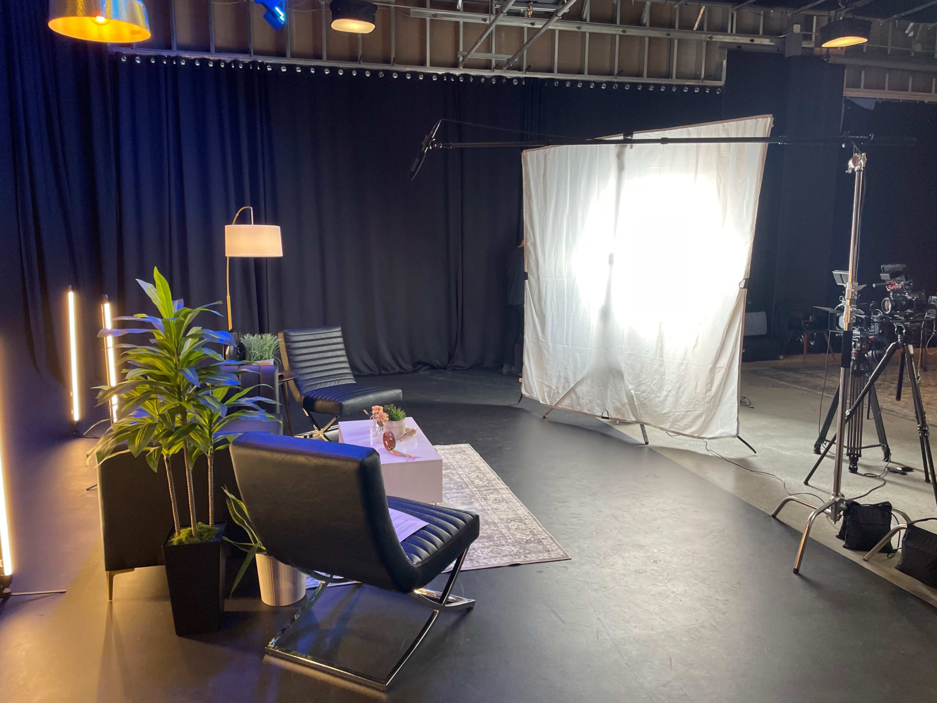 The image shows a studio setup with two black chairs, a small table with a decorative item, a standing lamp, and a large white diffusion panel to soften lighting in a backdrop of black curtains.