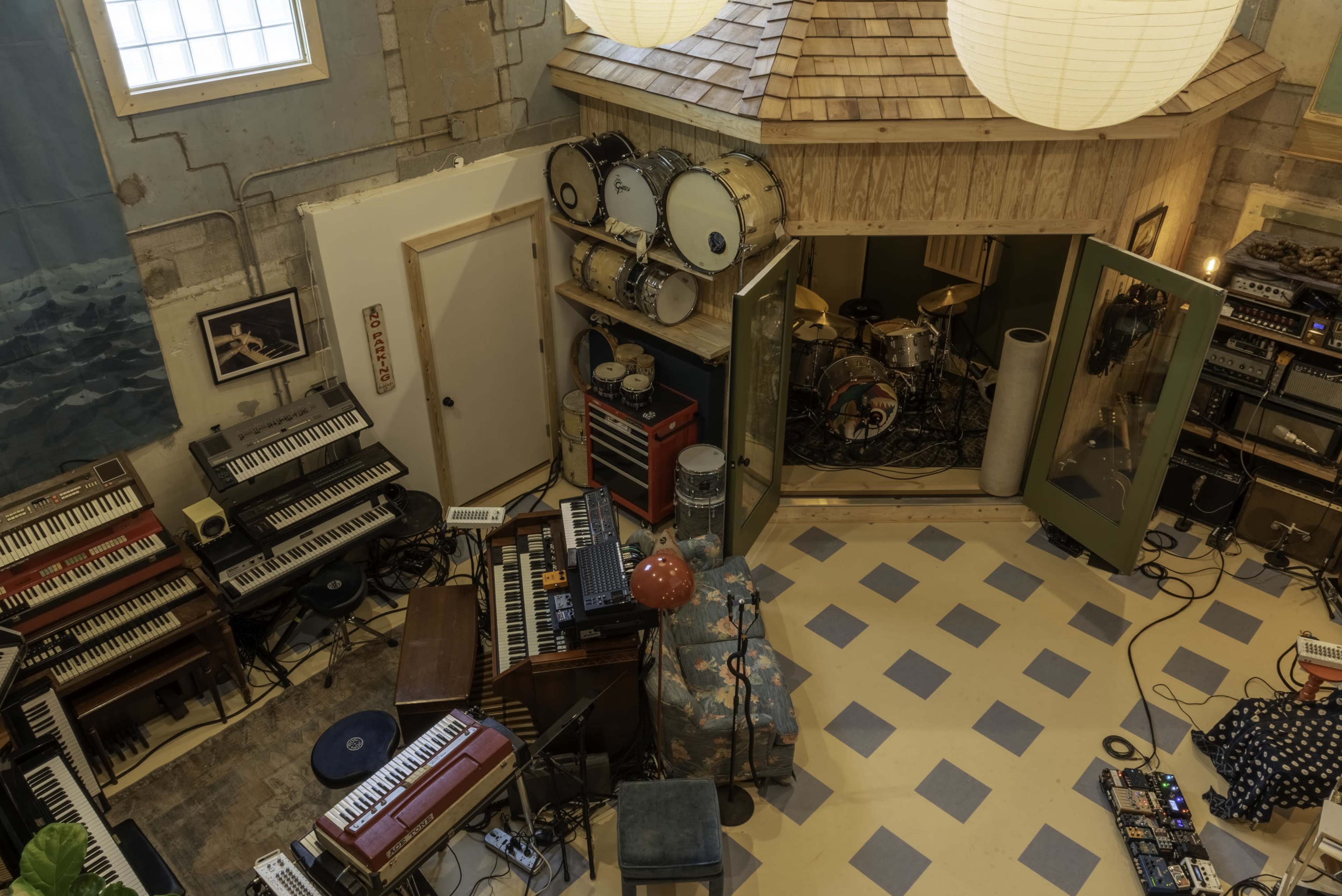 The image shows a music studio filled with various instruments, including keyboards, drums, and amplifiers, arranged in a spacious room with a unique roof structure.