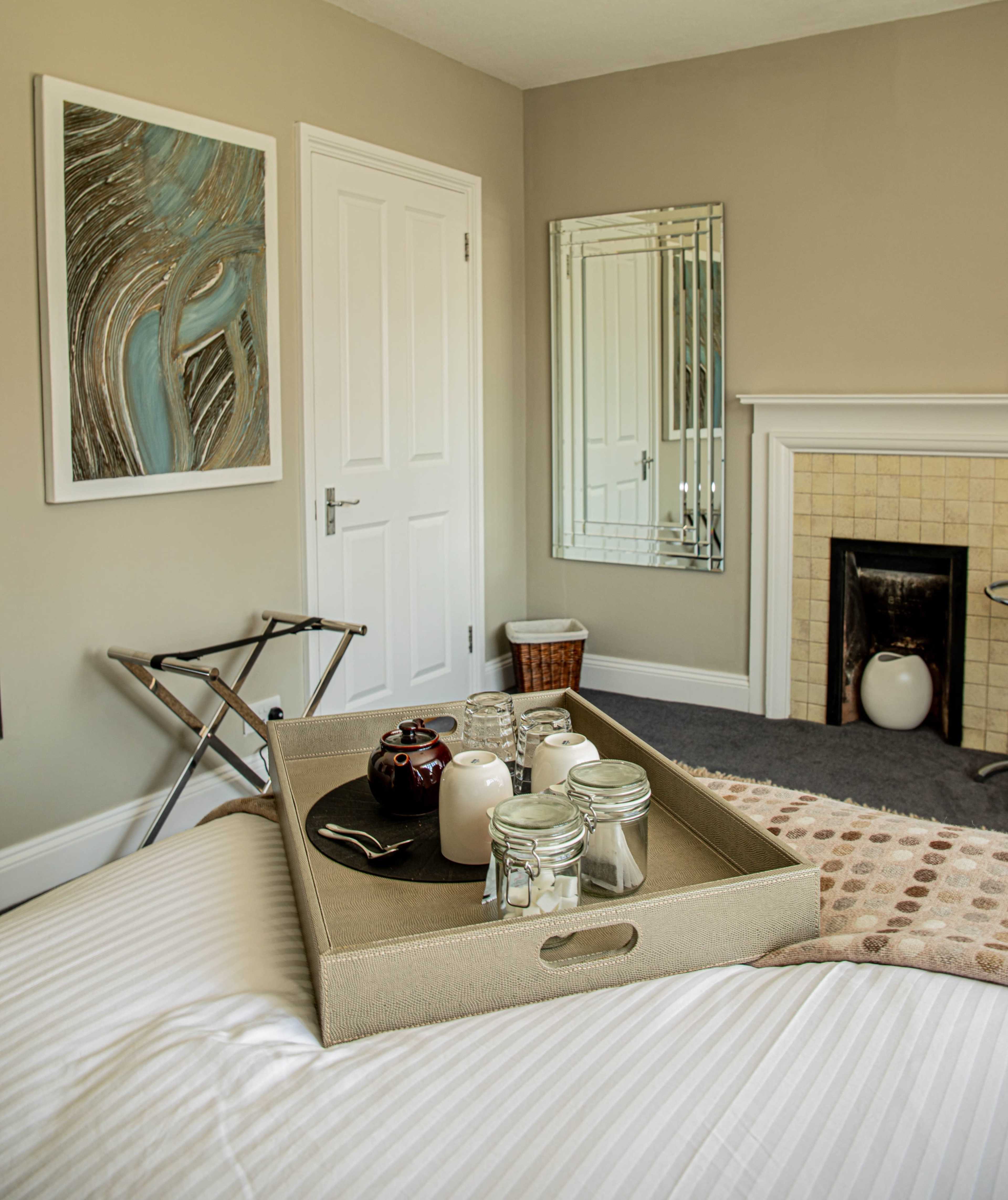 A neatly arranged bedroom features a bed with a tray of jars and a milk jug, while a mirror and artwork adorn the walls.