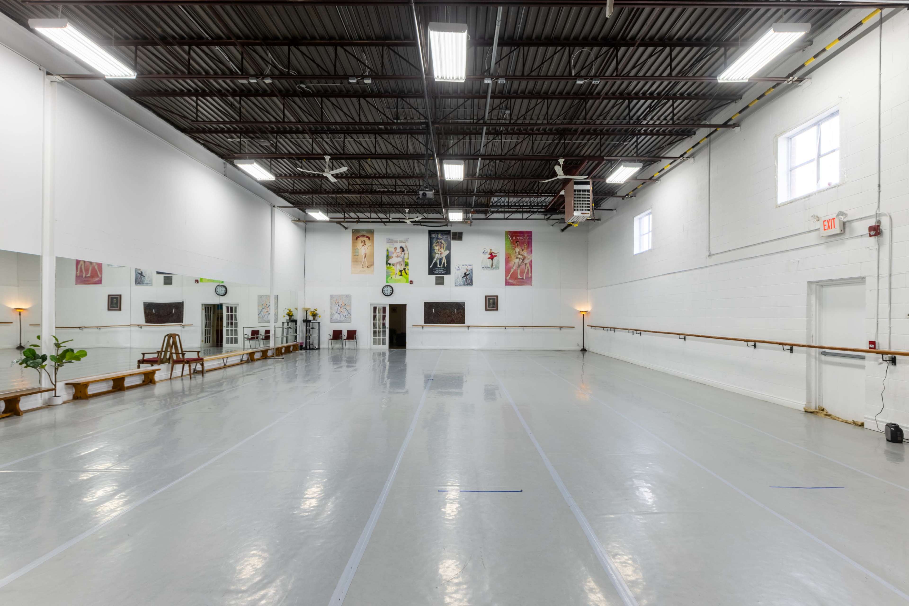 The image shows a spacious, empty dance studio with a mirrored wall, ballet barre, and bright lighting.