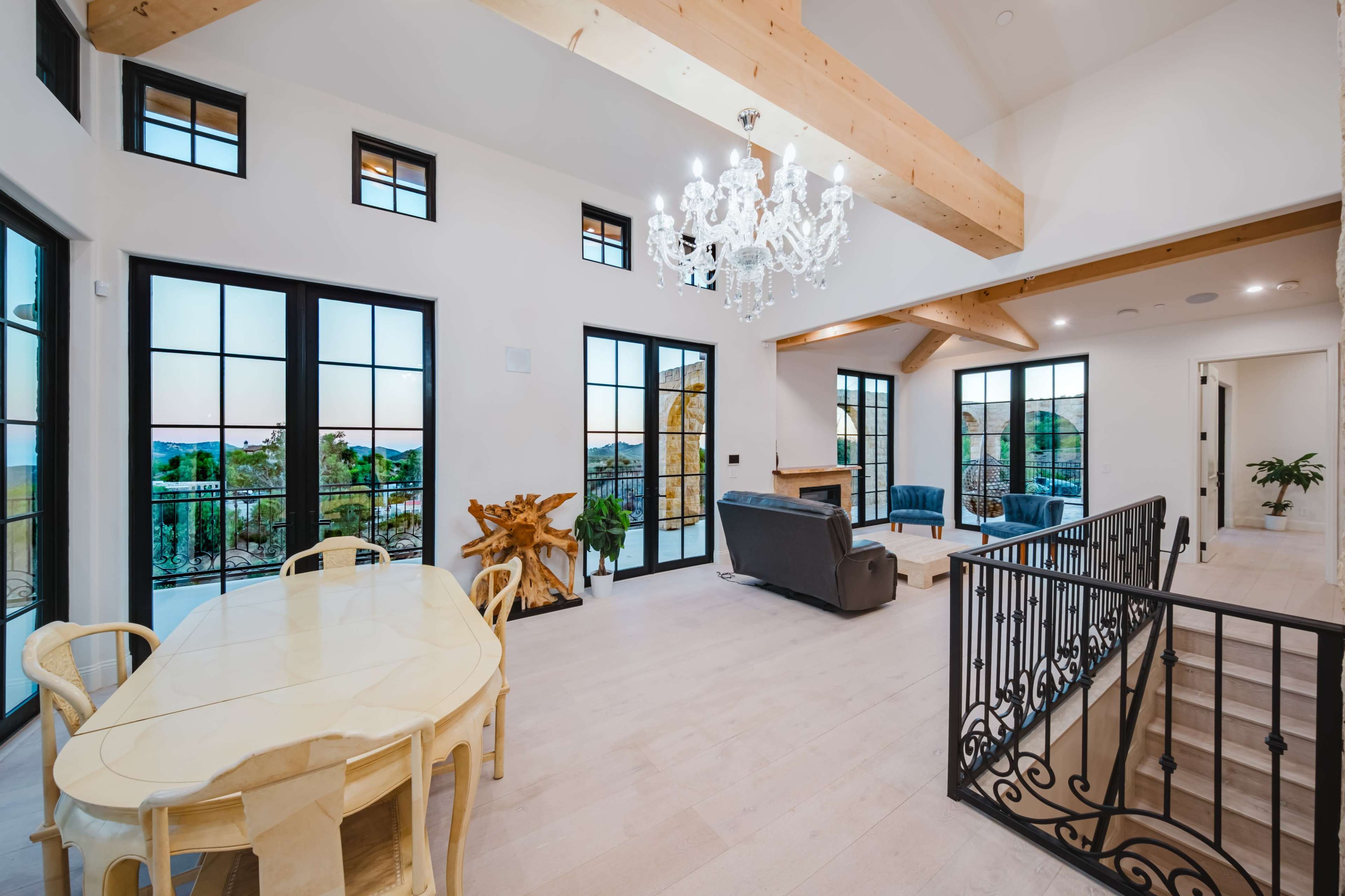 The image shows a modern open-concept living space with a dining area, a sitting area featuring a chandelier, large windows, and wooden beams.