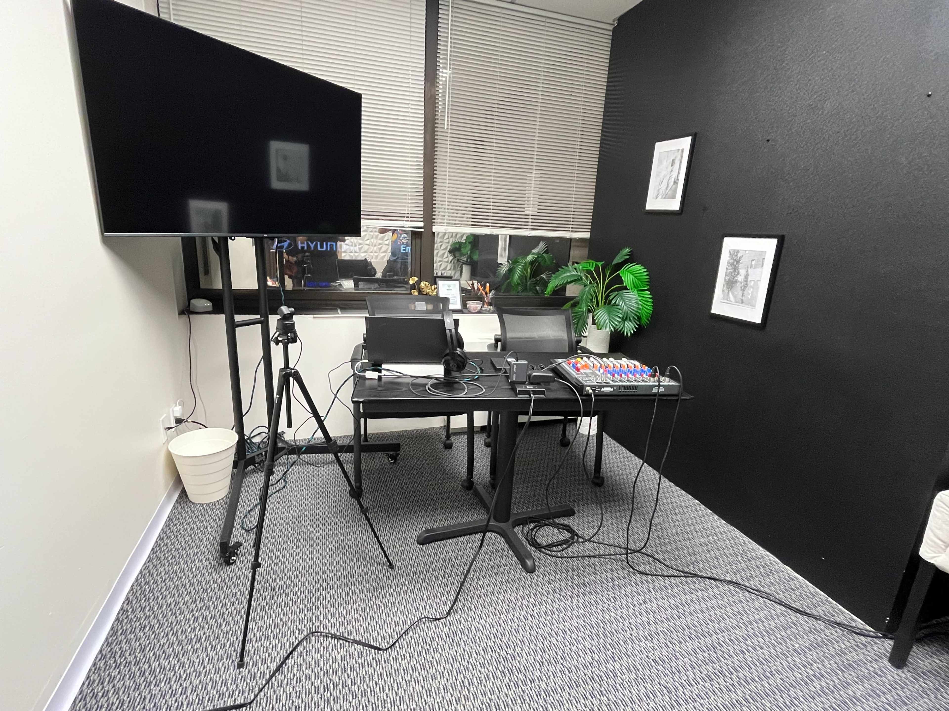 The image shows a small office space with two chairs, a table equipped with audio equipment, a television screen, and a few decorative items including a potted plant and framed artwork on the wall.