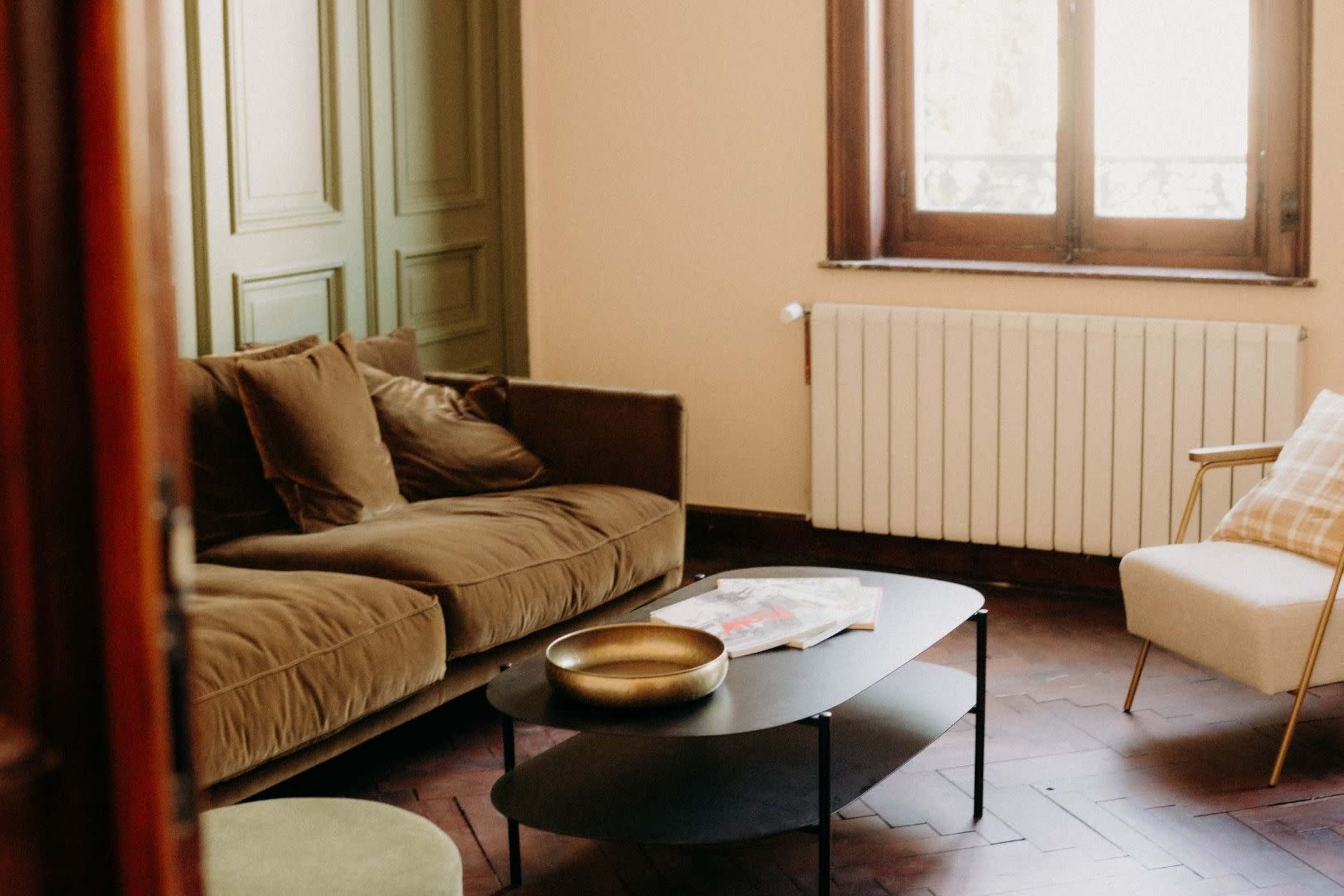 The image shows a cozy living room with a brown sofa, a round coffee table, and large windows allowing natural light to enter the space.