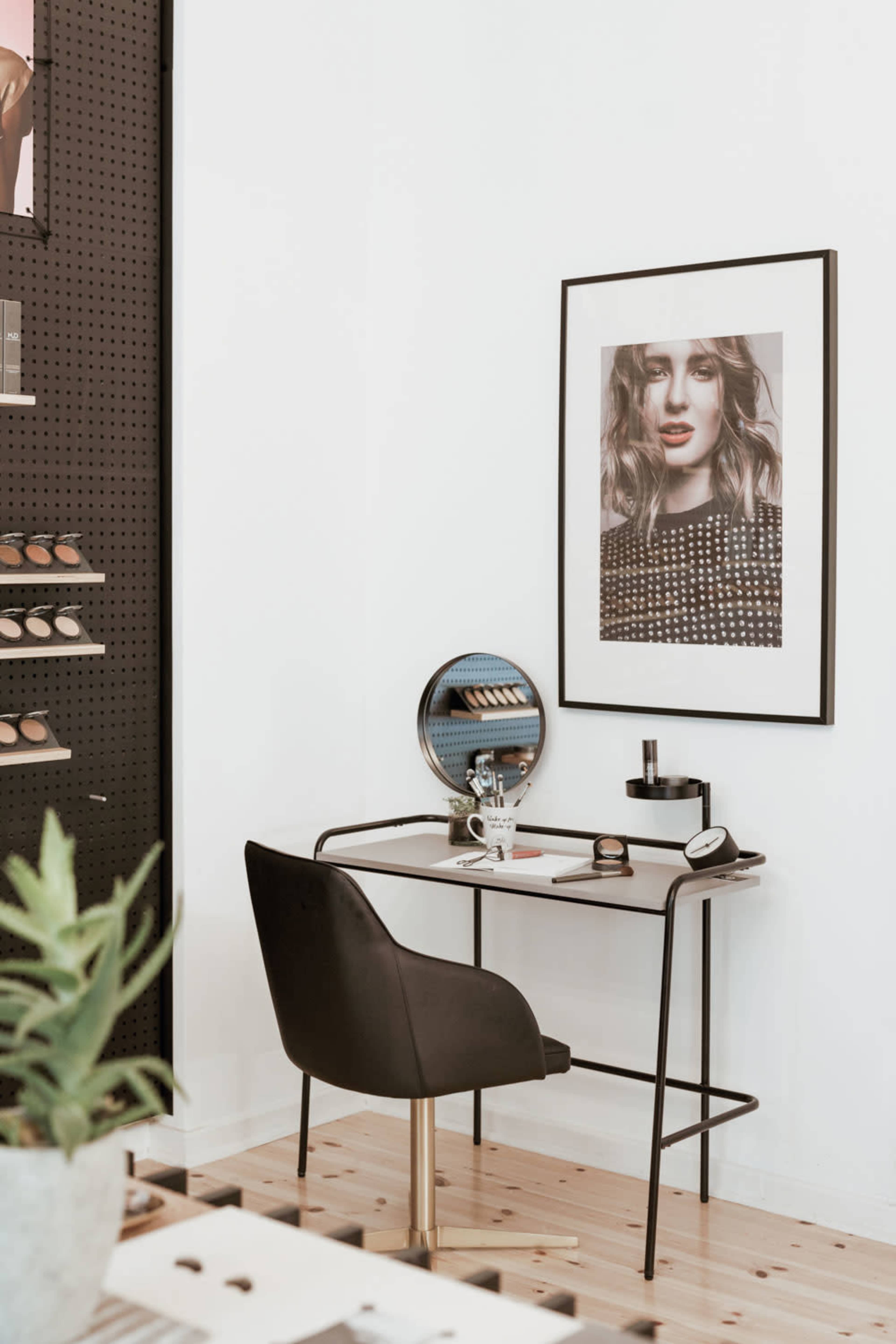 A minimalist vanity area features a chair, a round mirror, a desk with cosmetics, and a framed portrait on the wall.