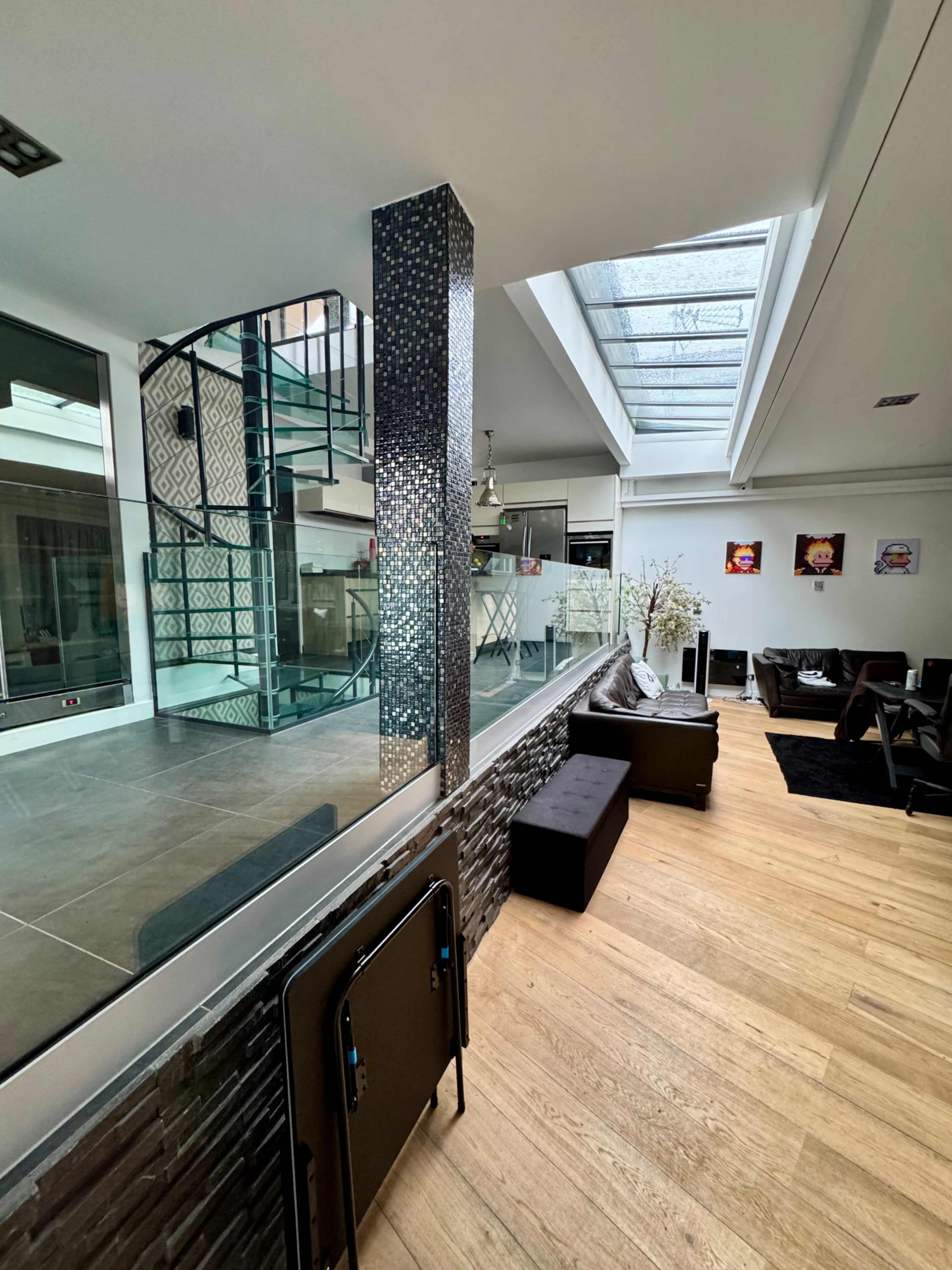 The image features a modern interior space with a glass staircase, a mix of dark and light furnishings, and large windows allowing natural light to enter.