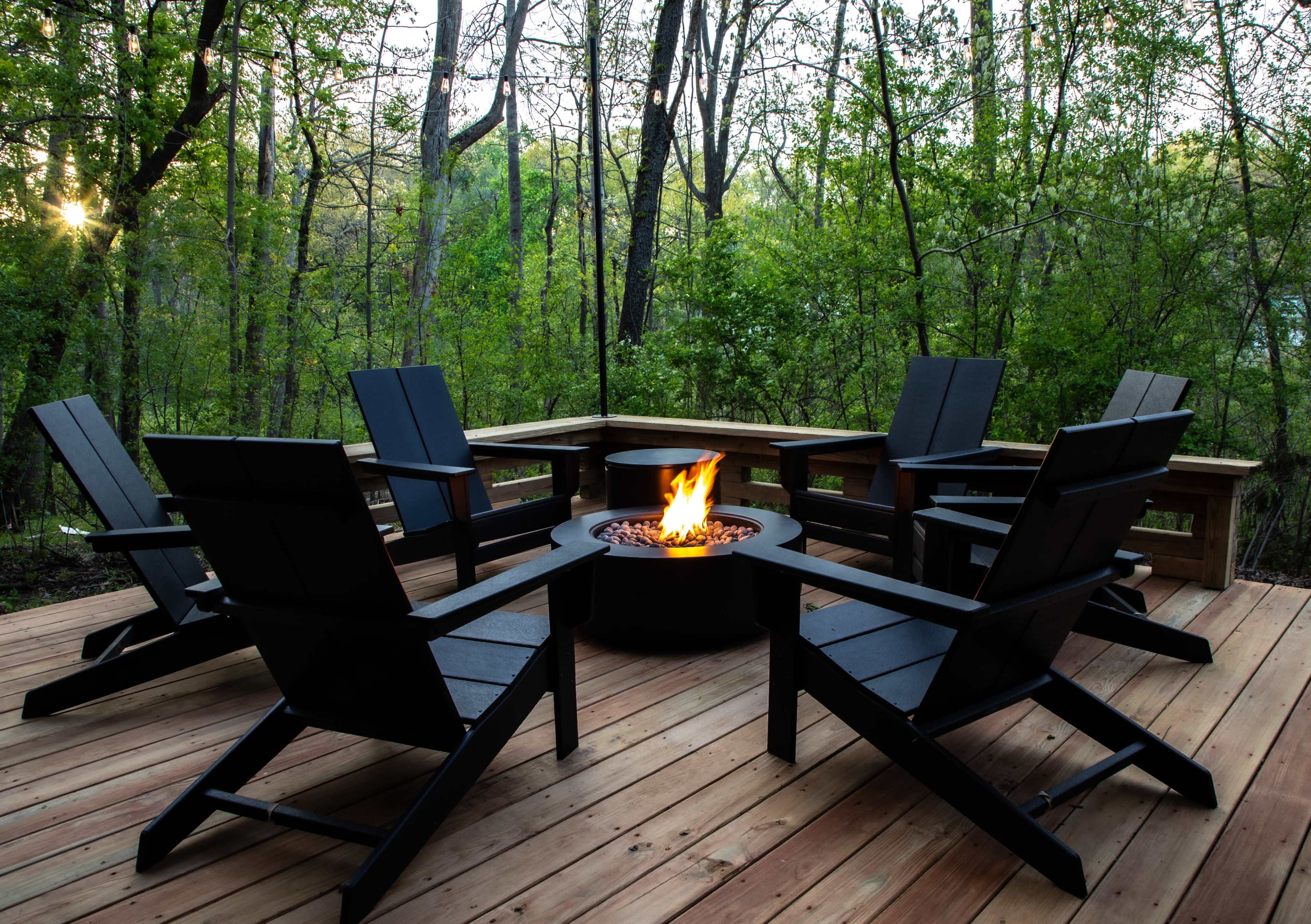 A circular arrangement of black Adirondack chairs surrounds a fire pit on a wooden deck, with greenery and trees in the background.