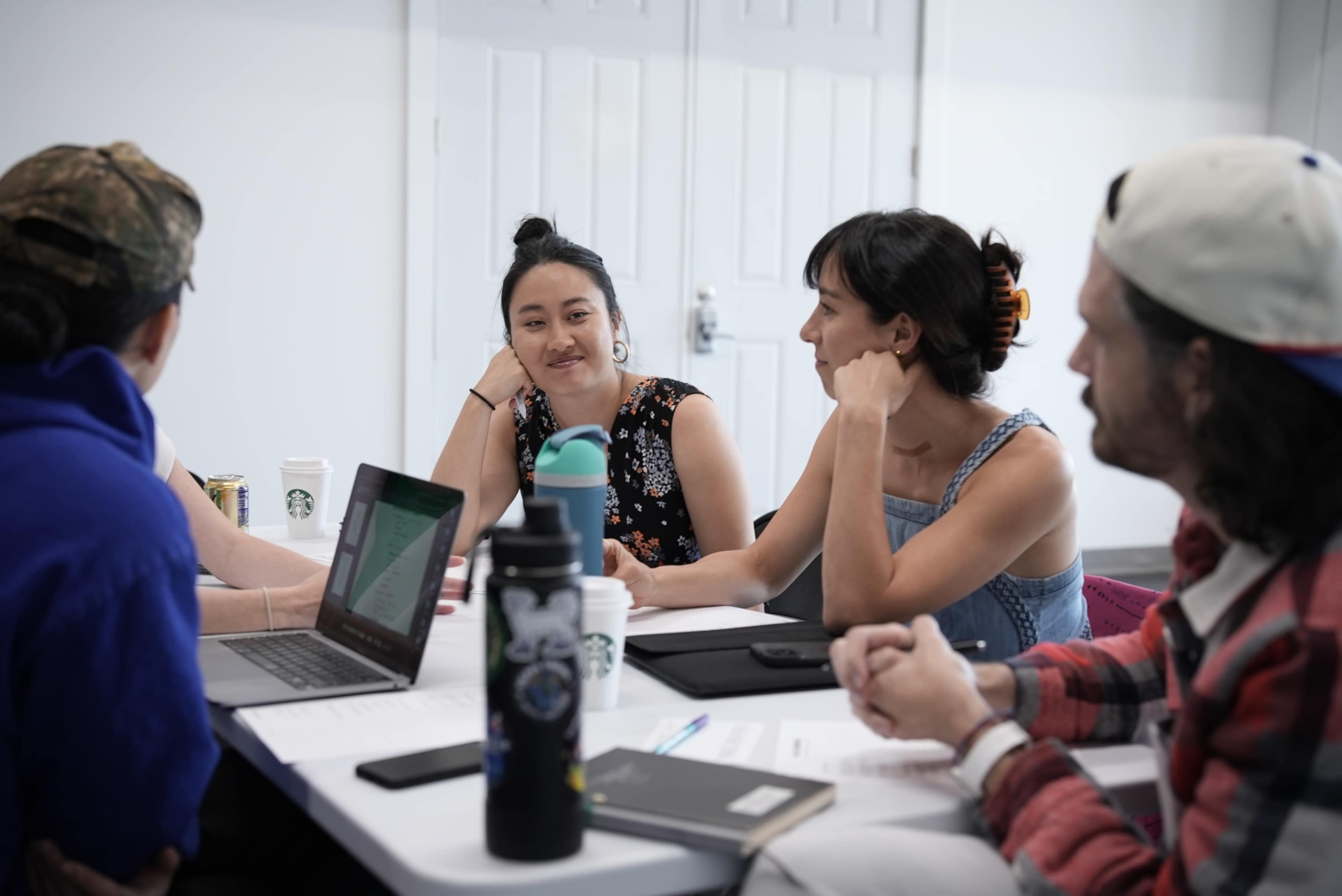 A group of five people engage in a discussion around a table with laptops and notebooks in a bright, modern room.