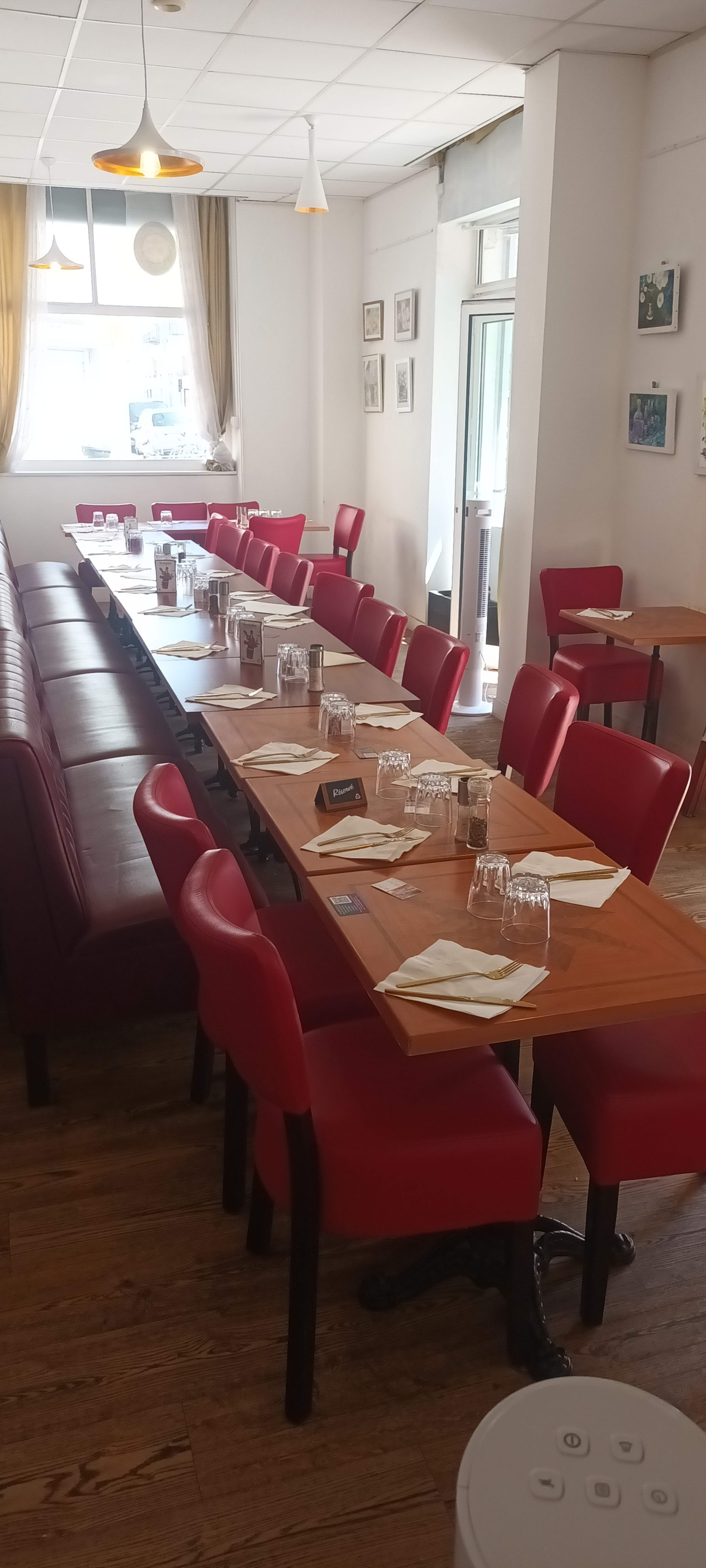 The image shows a bright restaurant interior with long tables arranged in the center, red chairs lining the tables, and a window letting in natural light.