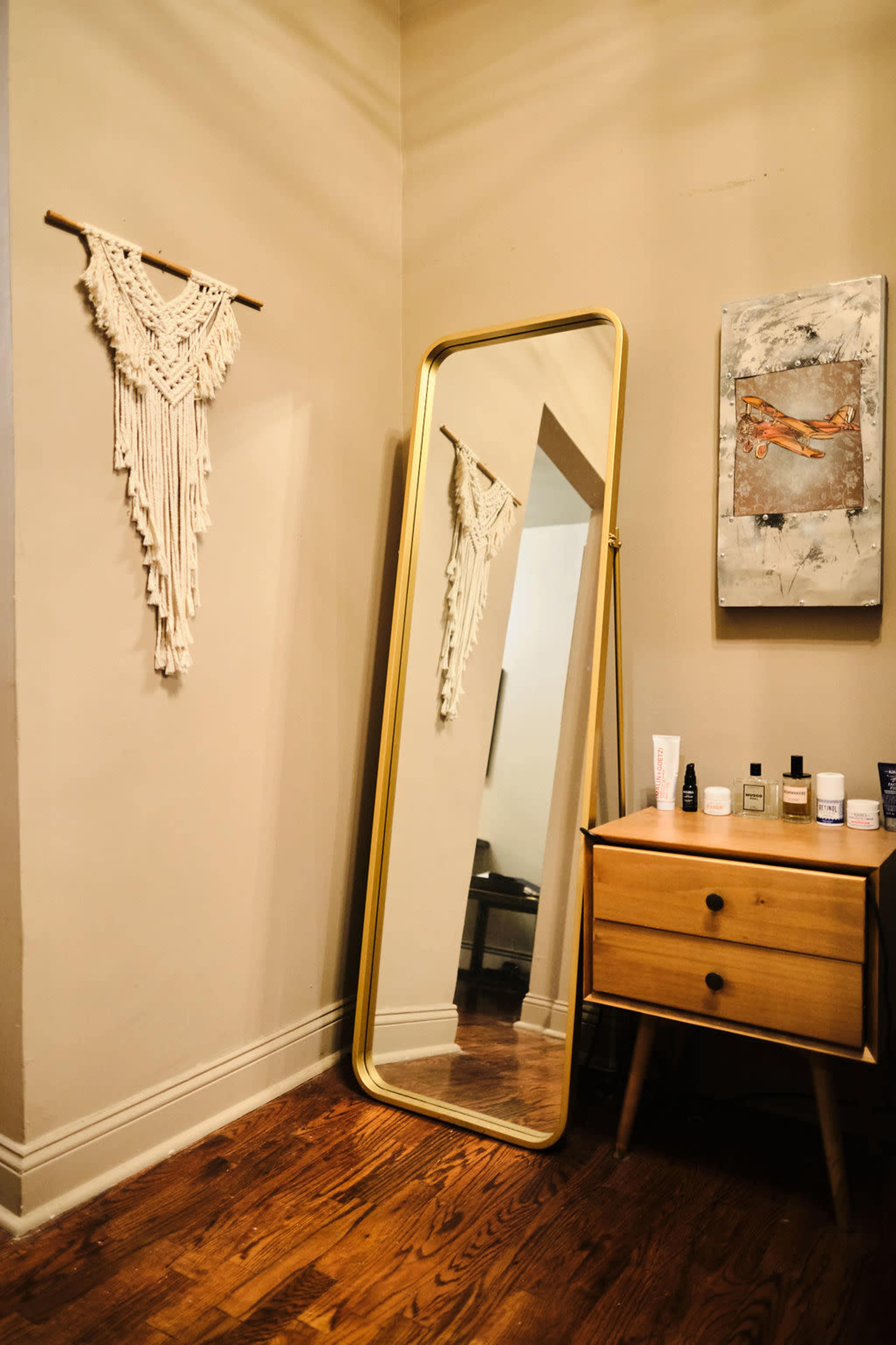A large gold-framed mirror leans against a wall next to a wooden dresser adorned with various skincare products and a wall hanging above it.