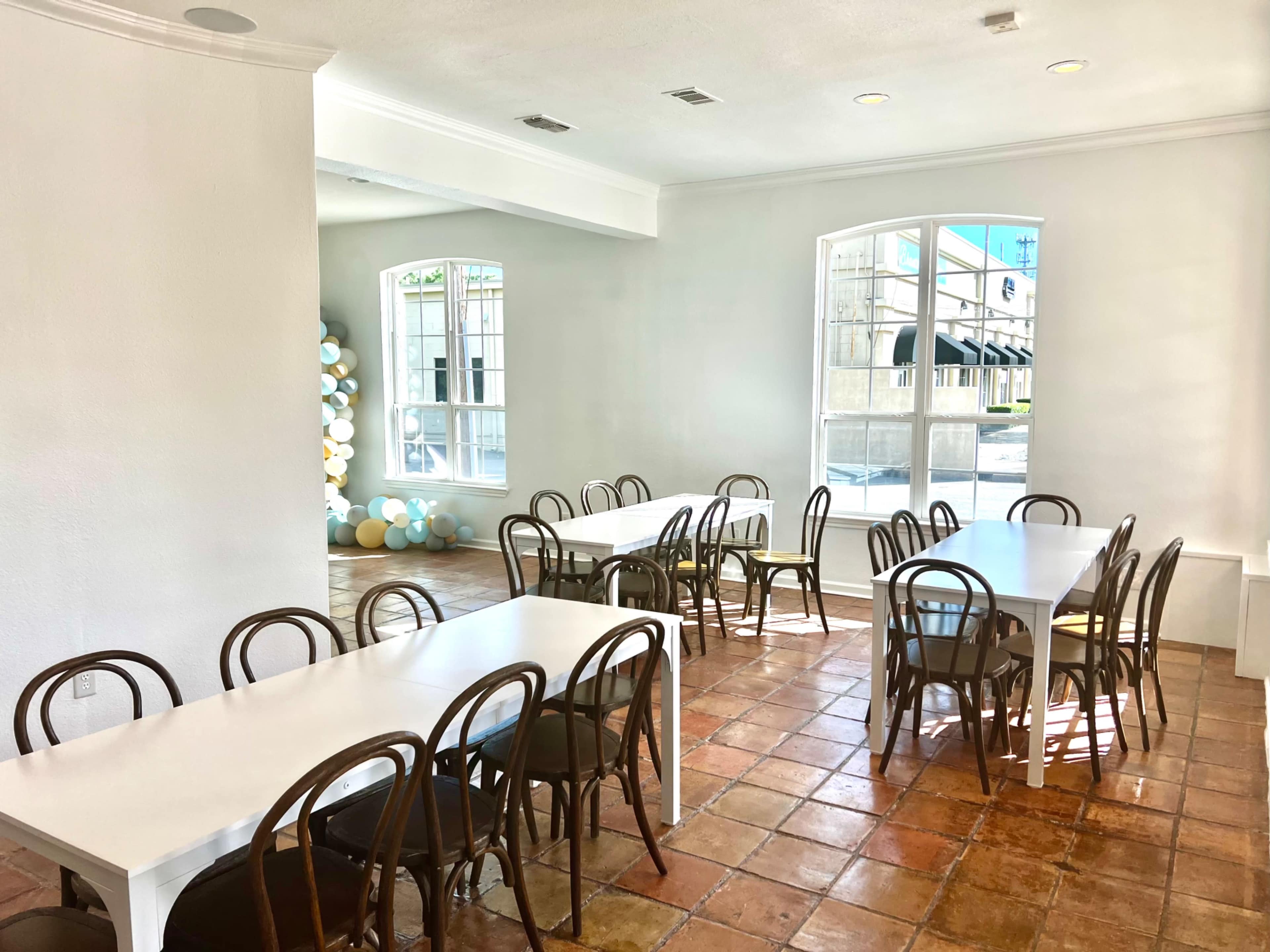 A bright room features several white tables and wooden chairs arranged on a tiled floor, with large windows letting in natural light.