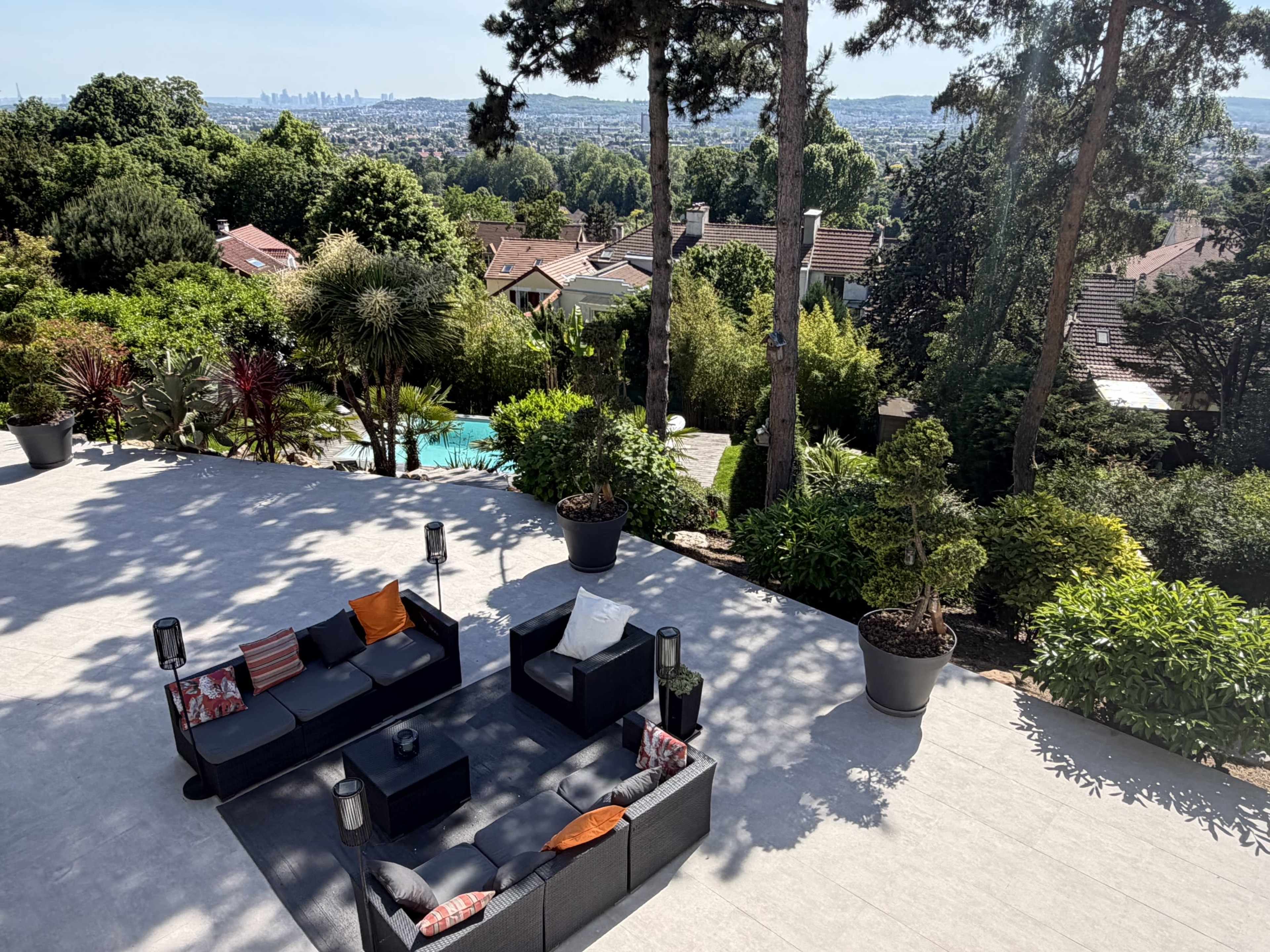 A modern patio with outdoor furniture overlooks a lush garden and a swimming pool, with a distant city skyline visible in the background.