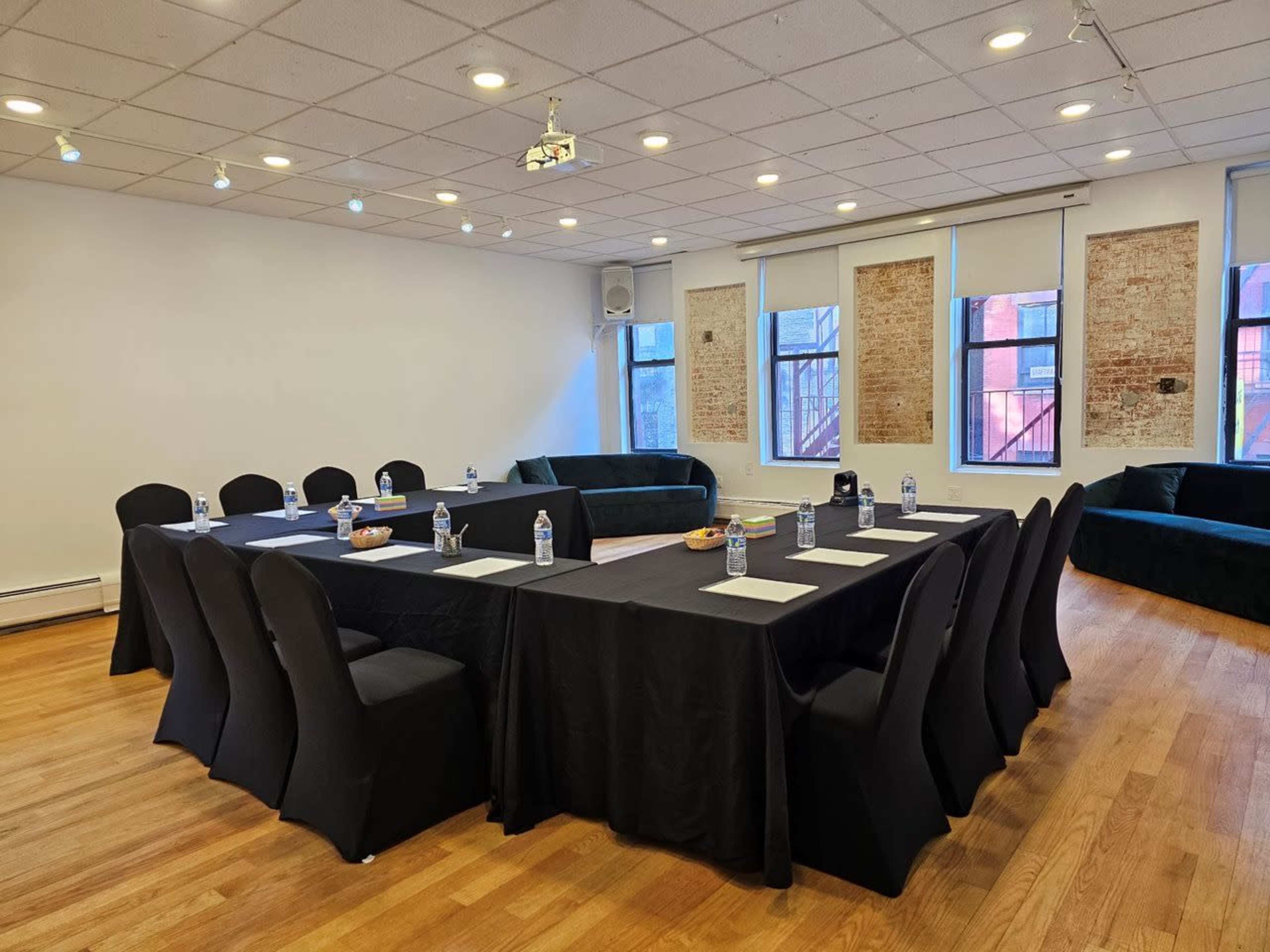 A conference room is set up with a long black table surrounded by black chairs, water bottles, and notepads, alongside a green couch against a brick wall.