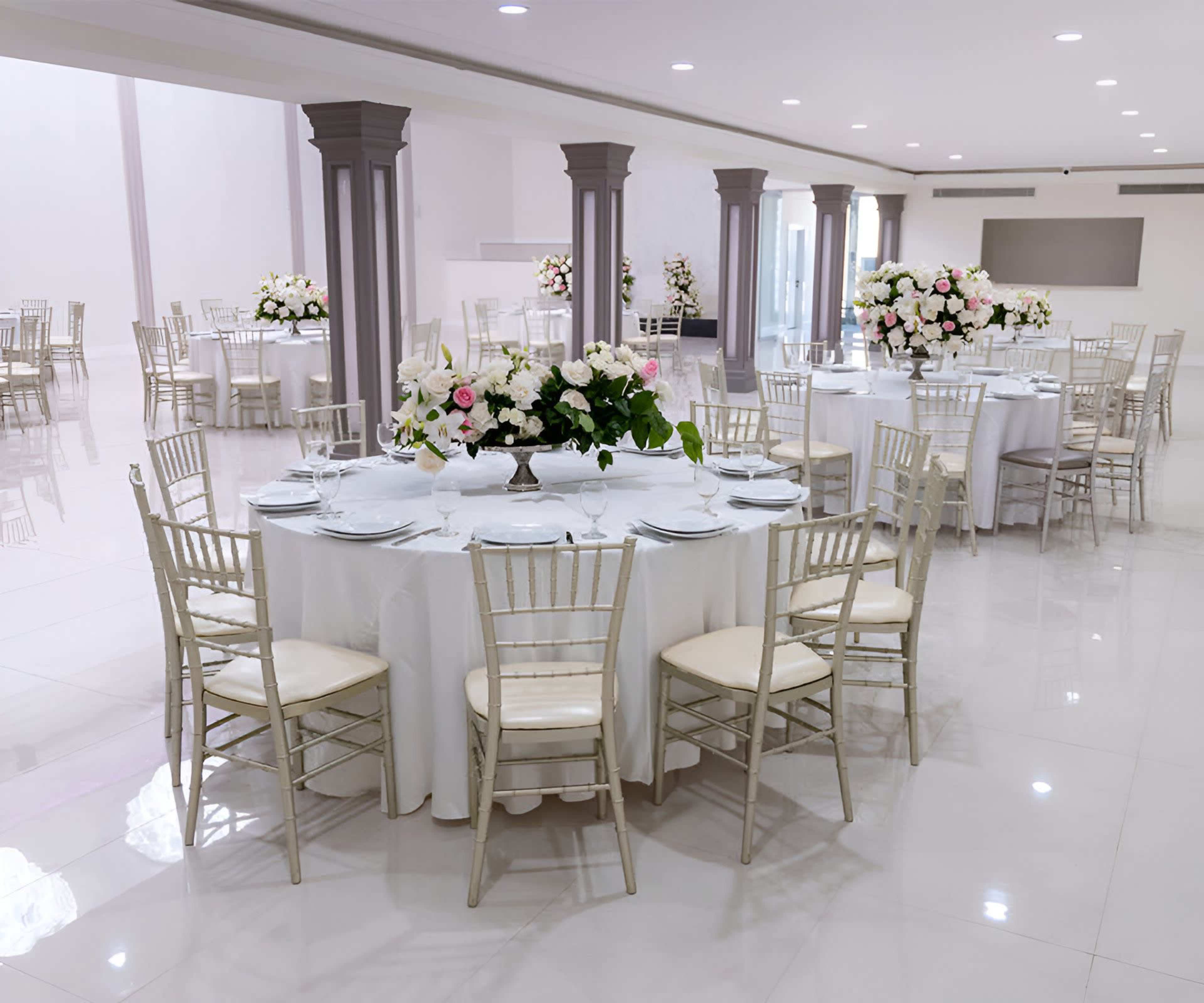 A spacious banquet hall is set up with round tables covered in white tablecloths and adorned with floral arrangements.