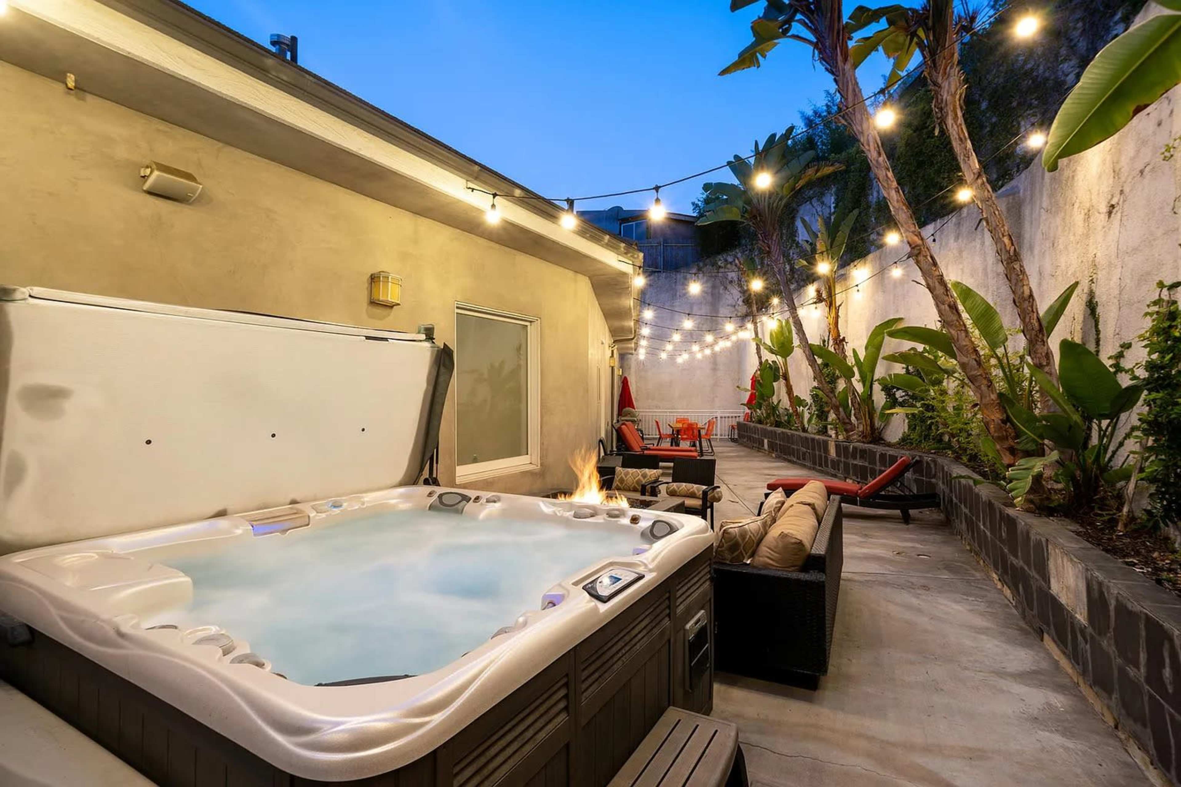 A backyard space featuring a hot tub surrounded by potted plants, outdoor seating, and string lights overhead.