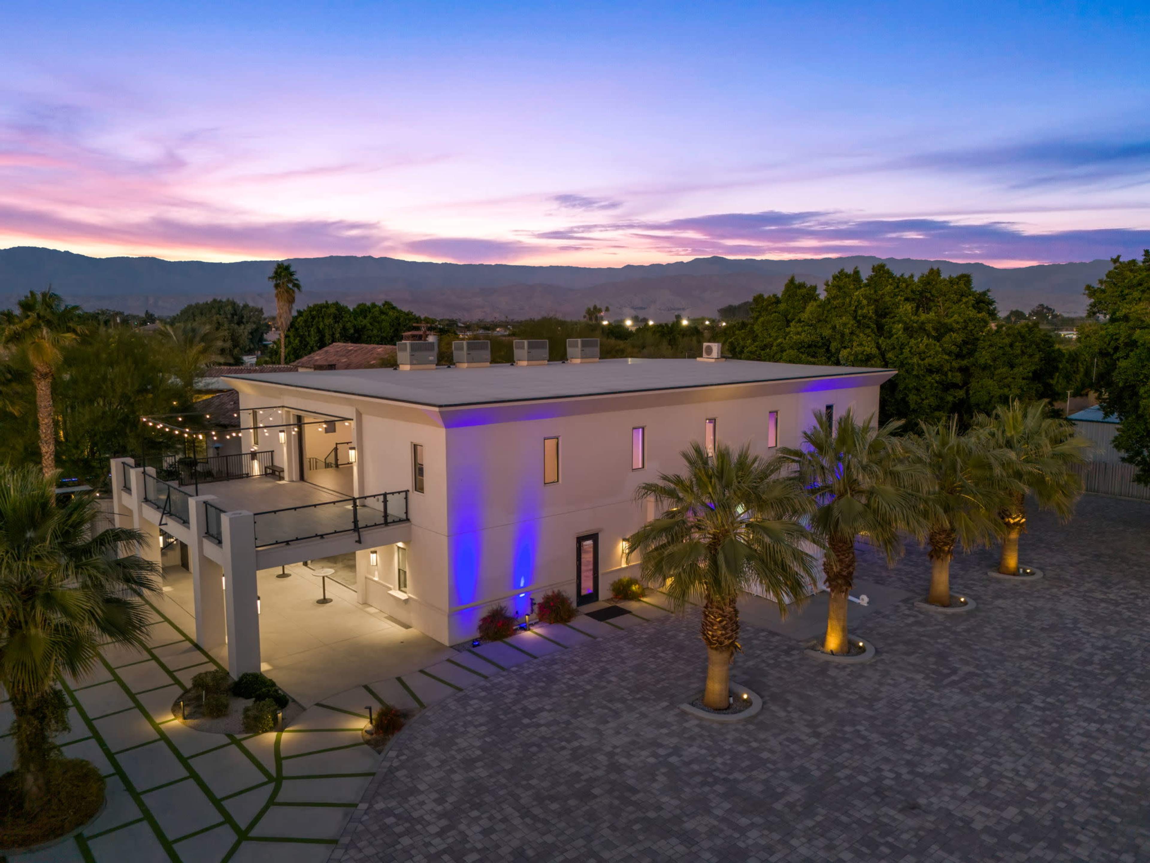 A modern two-story house with palm trees and a paved driveway is illuminated by soft lights against a twilight sky.