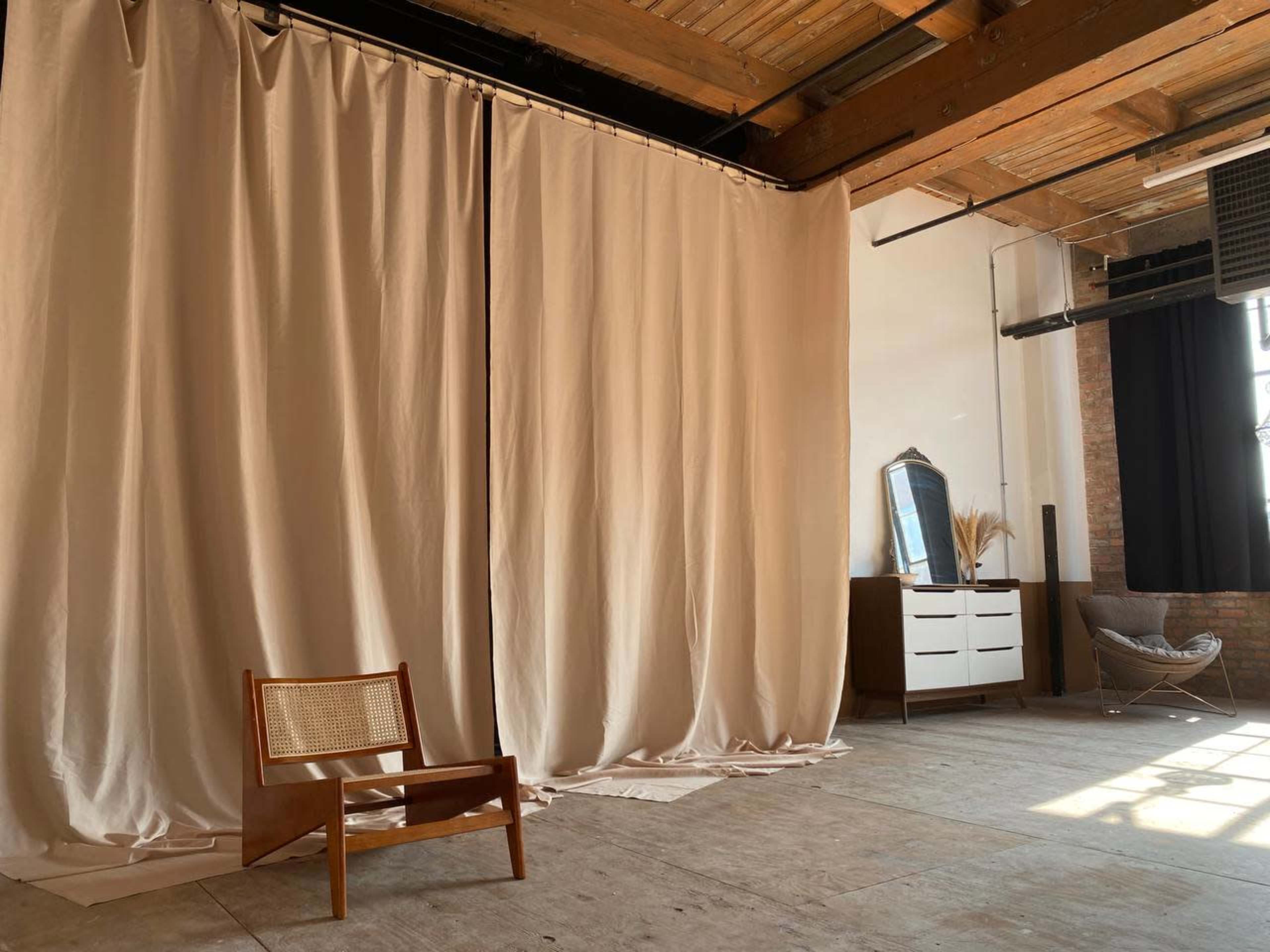 The image shows a spacious interior with wooden beams, large fabric curtains, a mid-century modern chair, a dresser, and a mirror against a brick wall.