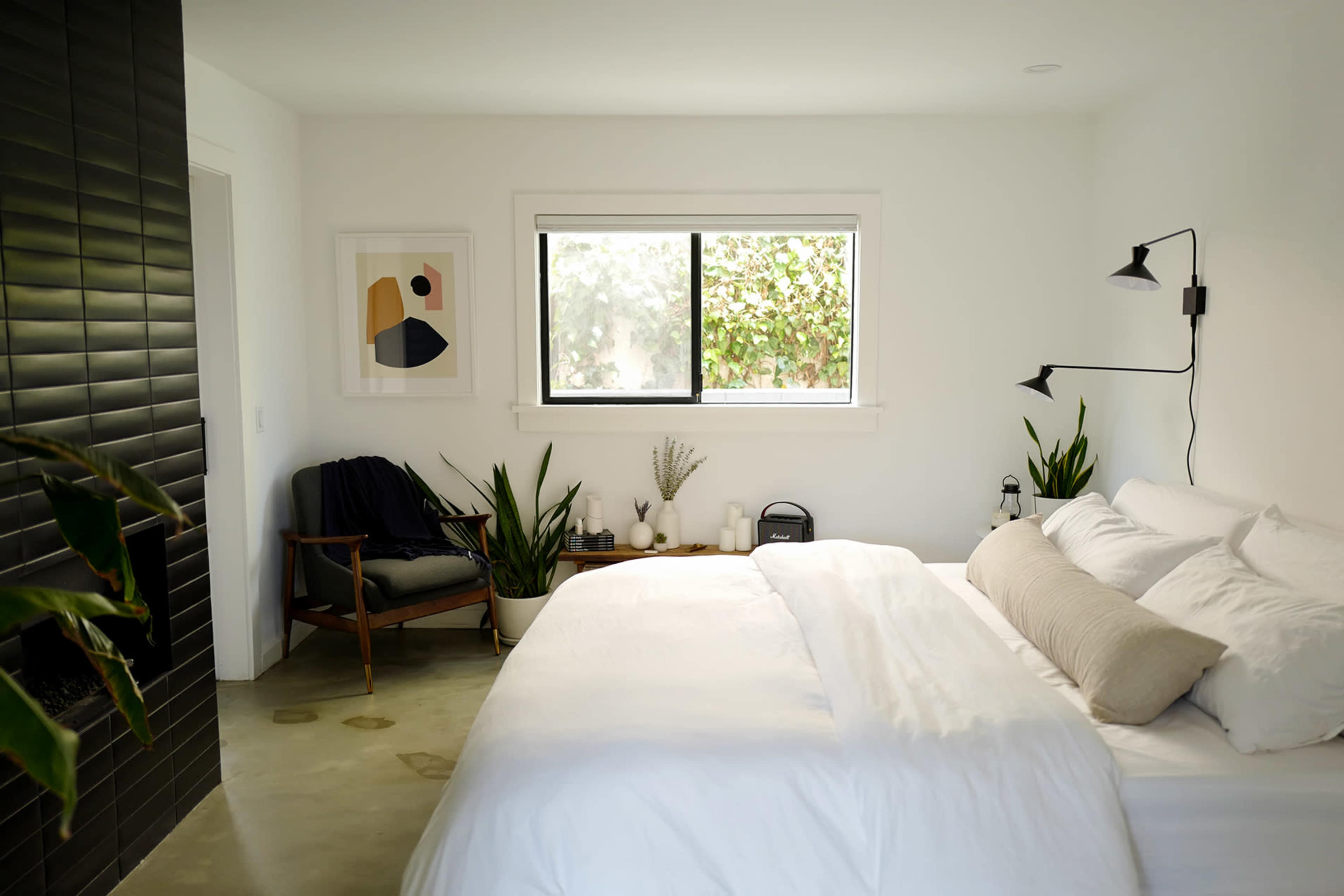 A minimalist bedroom features a large bed with white bedding, a chair beside the window, and several plants arranged throughout the room.