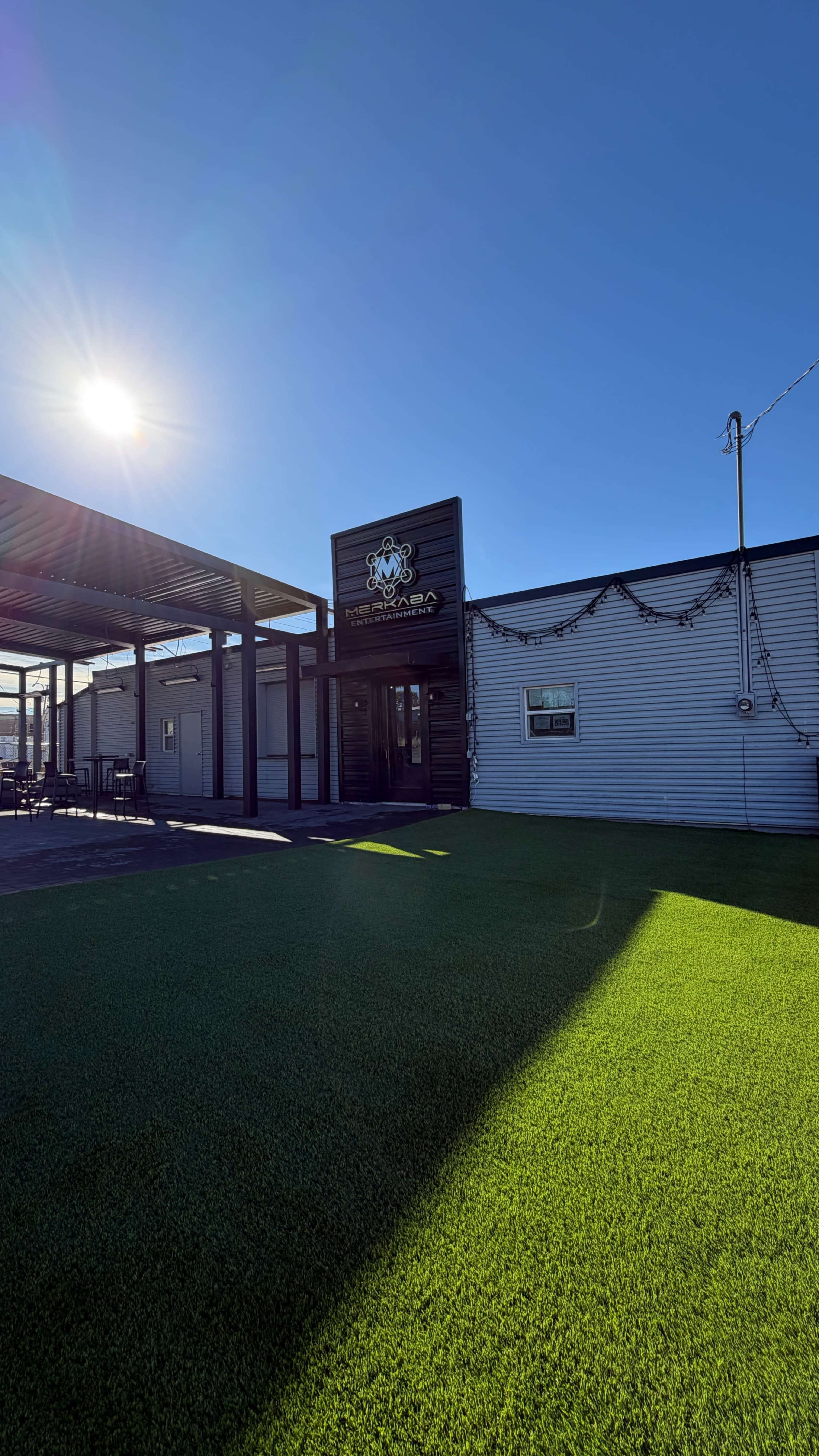The image shows a modern building with a sign featuring a logo, surrounded by green artificial grass, under a clear blue sky with sunlight shining.