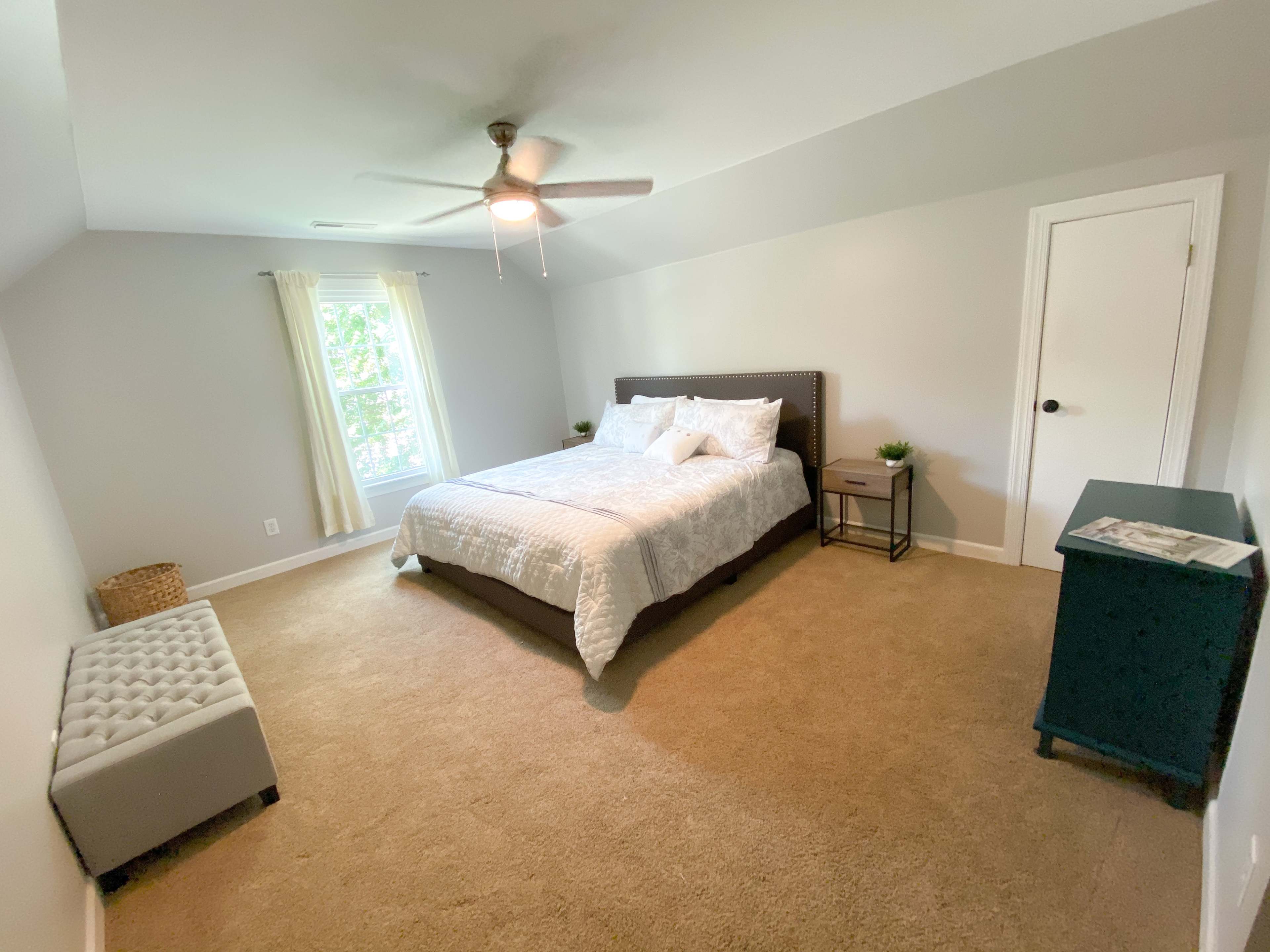 A spacious bedroom with a large bed, a nightstand, a small storage bench, and a window allowing natural light to enter.