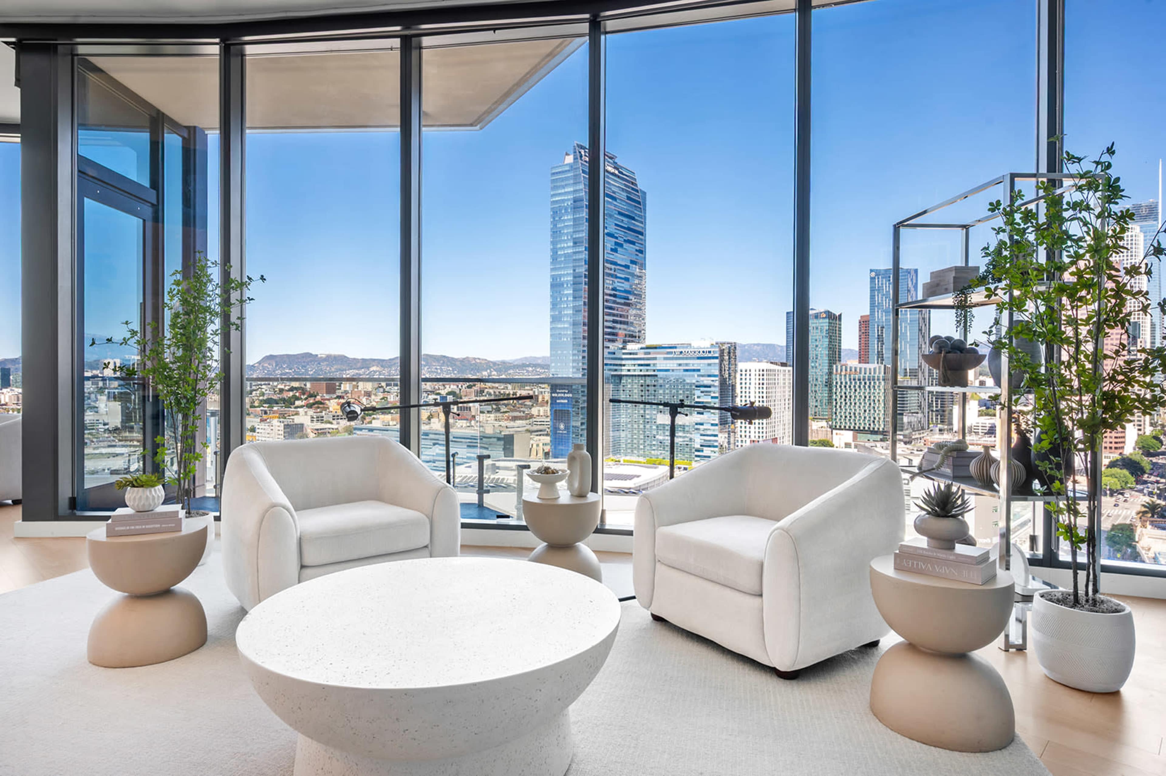 The image shows a modern living space with two white chairs and a round coffee table, set against a large window offering a view of a city skyline.