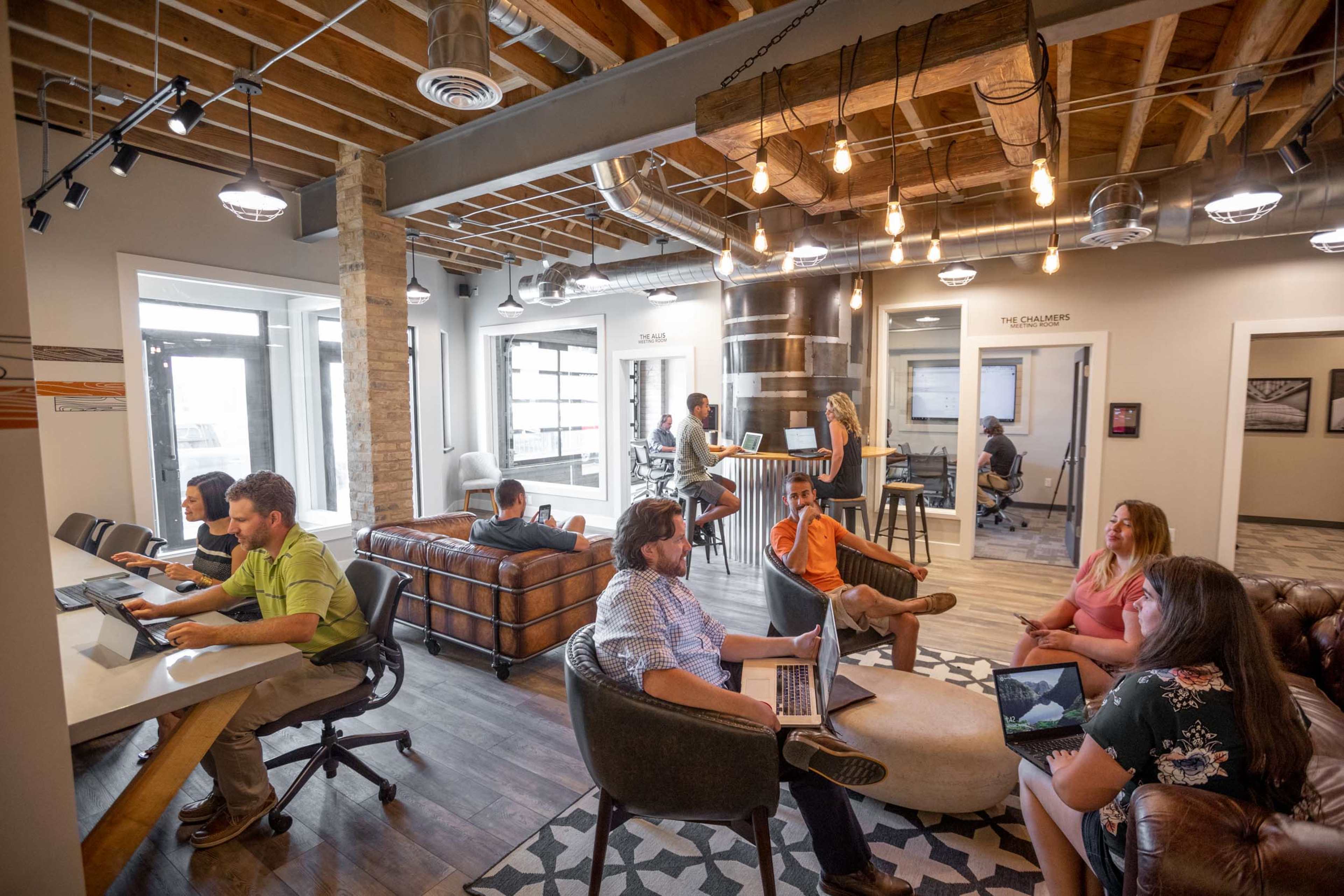 A modern co-working space features people engaged in various activities, with some working on laptops and others conversing in a comfortable, open layout.