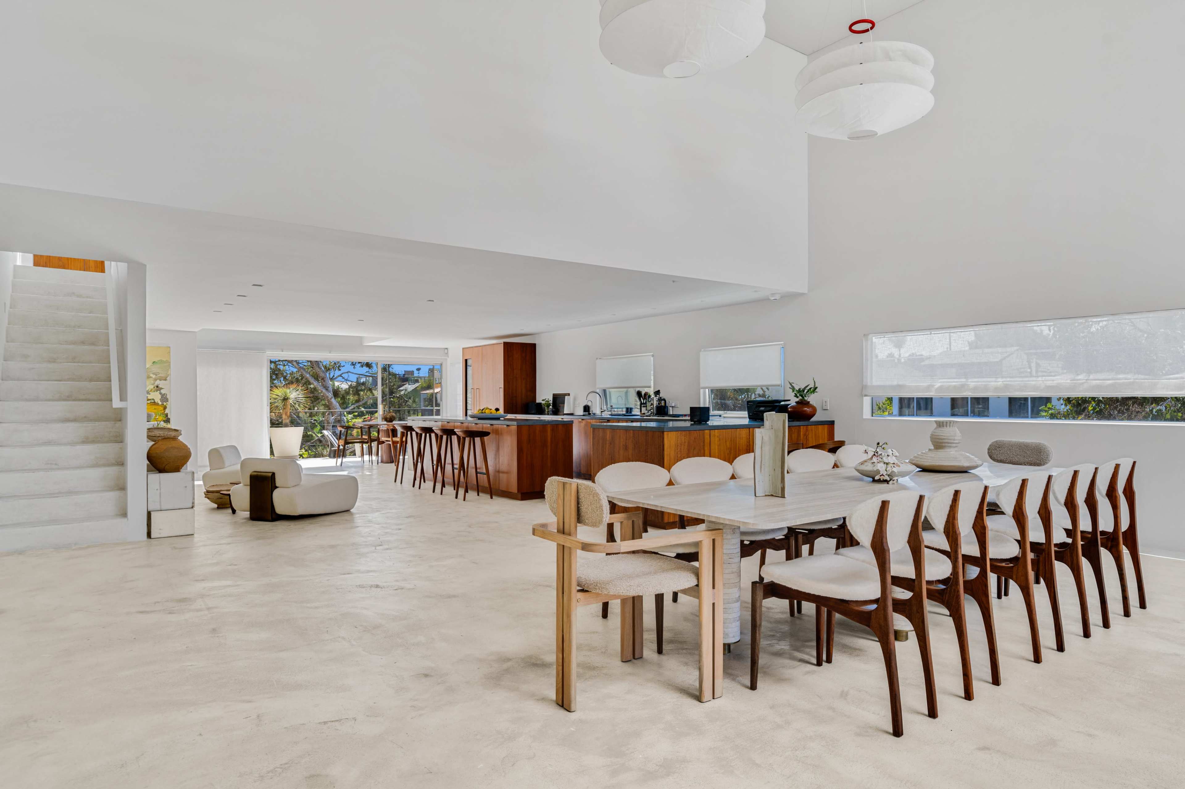 The image shows a modern, open-plan dining area featuring a long wooden table with wooden chairs, a kitchen bar in the background, and a staircase on the left.