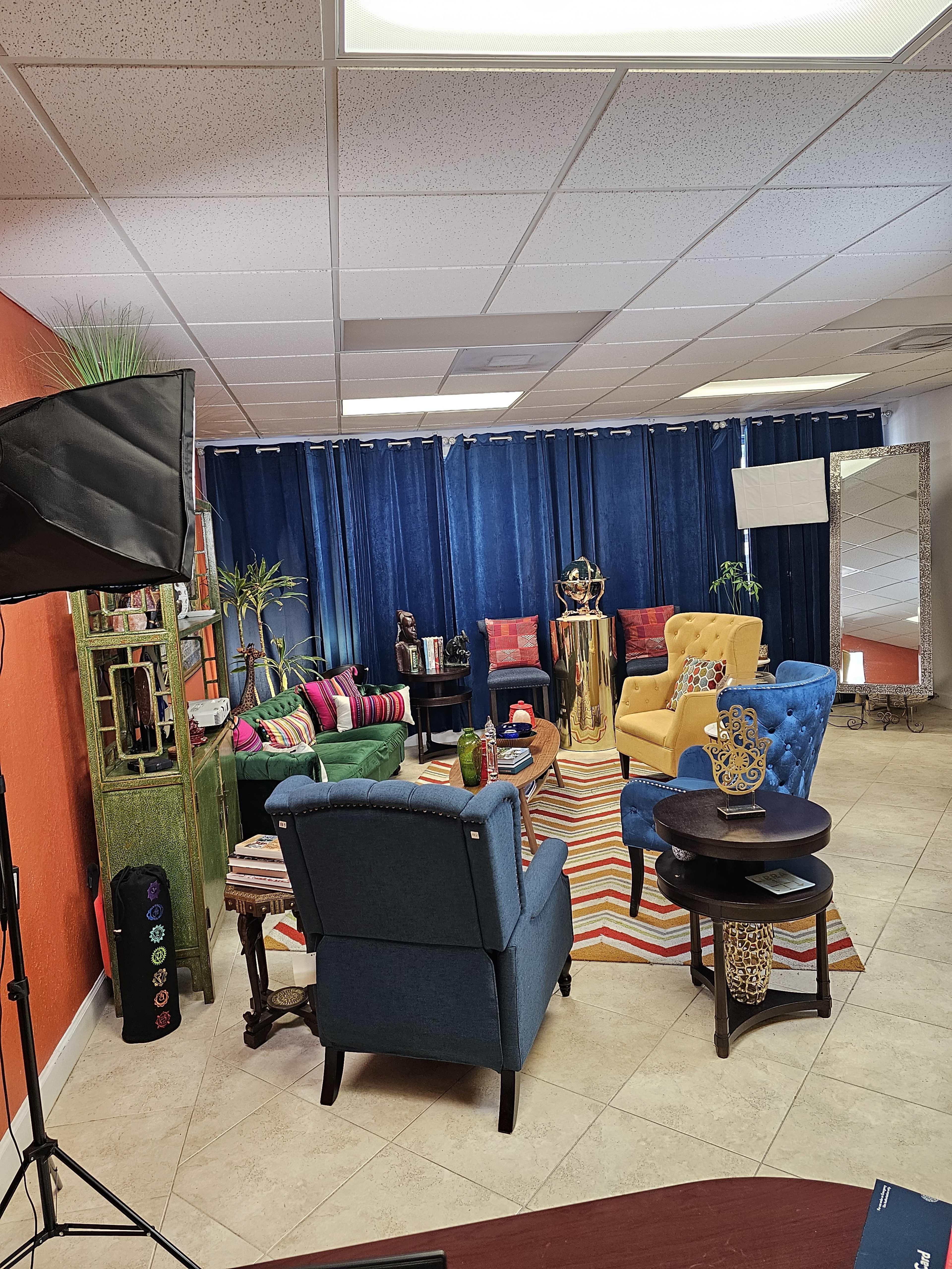 The image shows a colorful interior seating area with various chairs and a table, surrounded by curtains, plants, and decorative items.