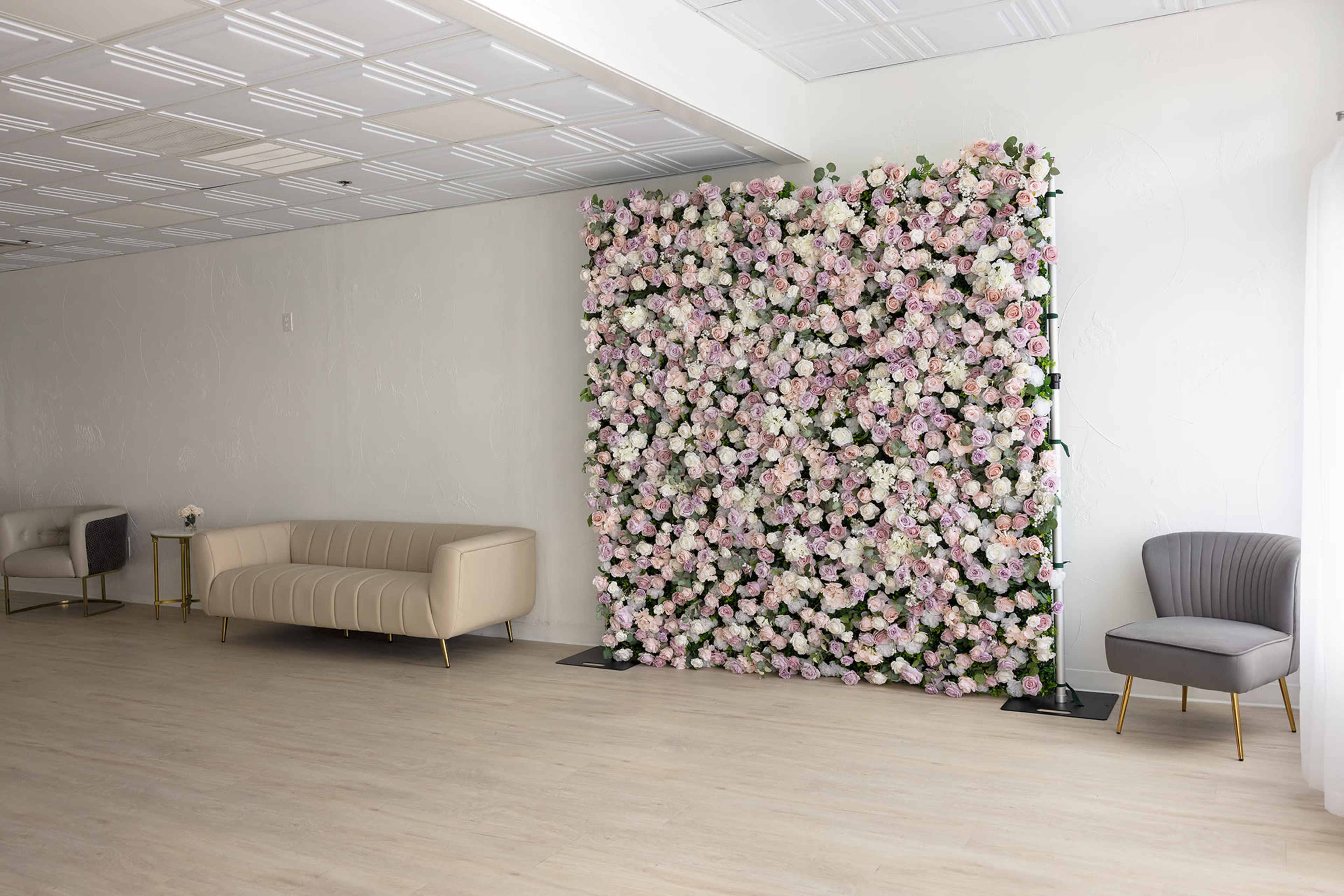 The image shows a large floral wall made of pink and white flowers, set against a light-colored wall in a spacious room with minimal furniture.