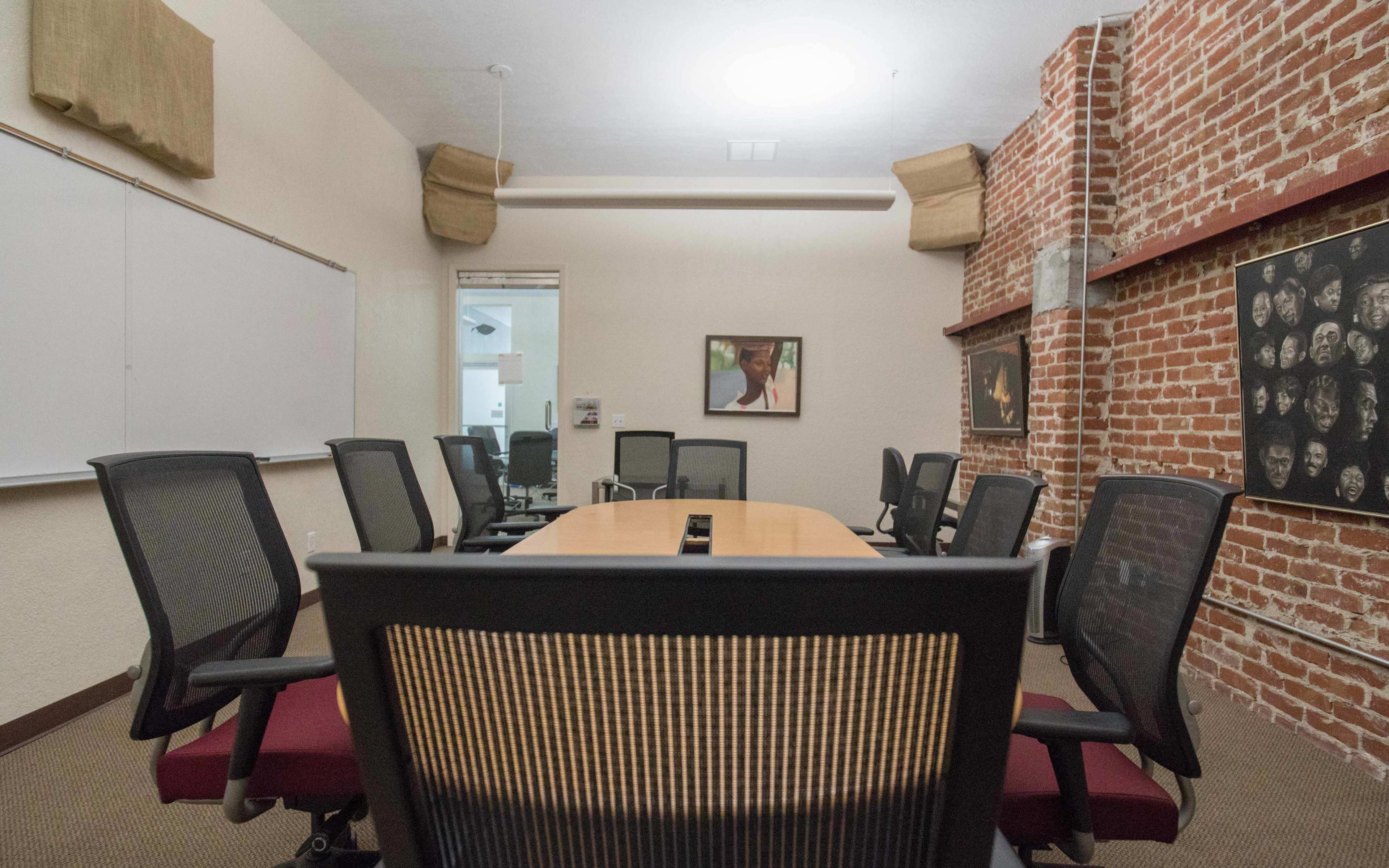 A meeting room with a long wooden table surrounded by black mesh chairs and exposed brick walls, featuring framed artwork on the walls.