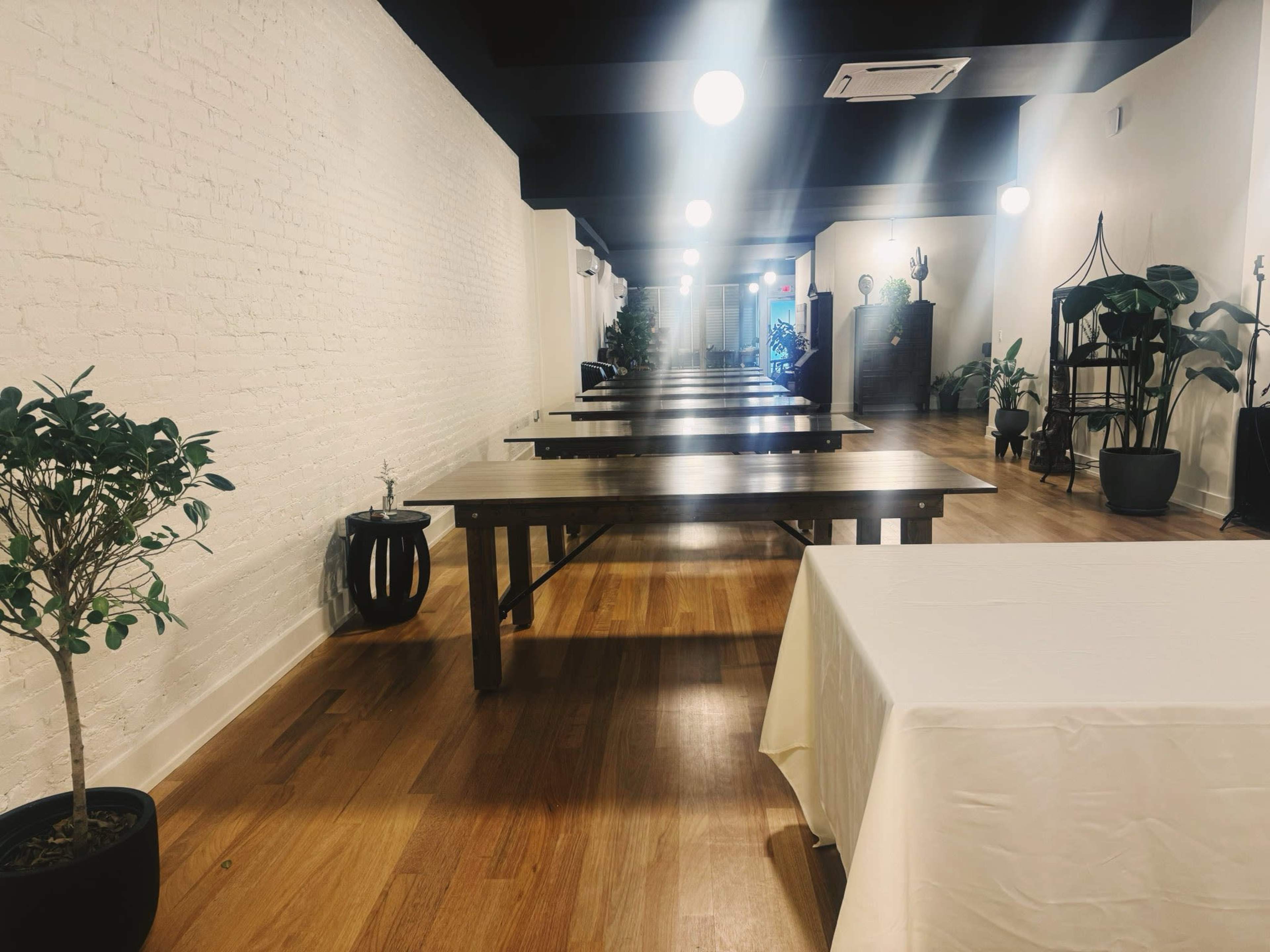 A long, empty dining area features several wooden tables lined up under hanging lights, with potted plants along the walls.
