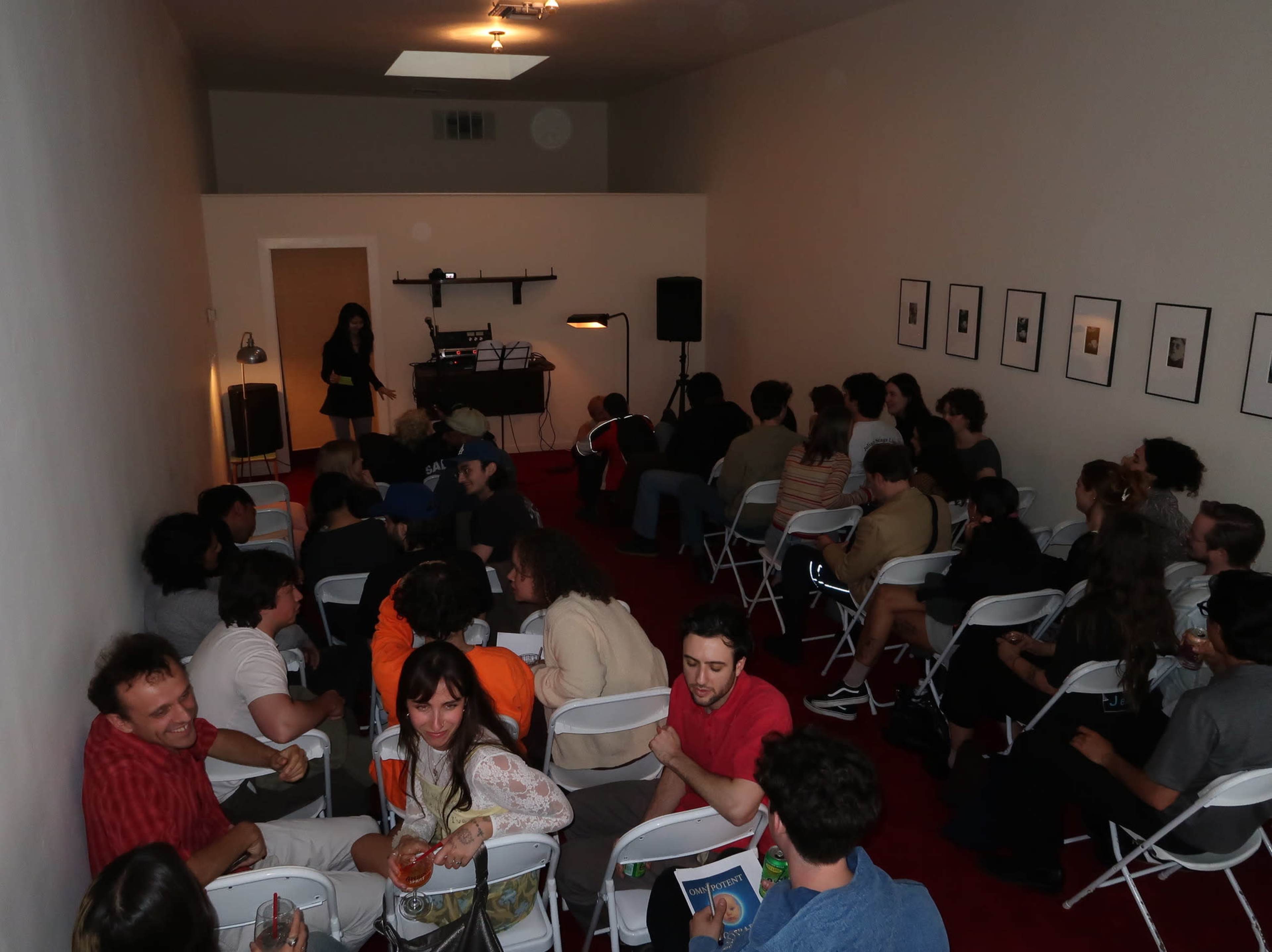 A crowded room with rows of chairs facing a small stage where a person is speaking, while others are seated and socializing.