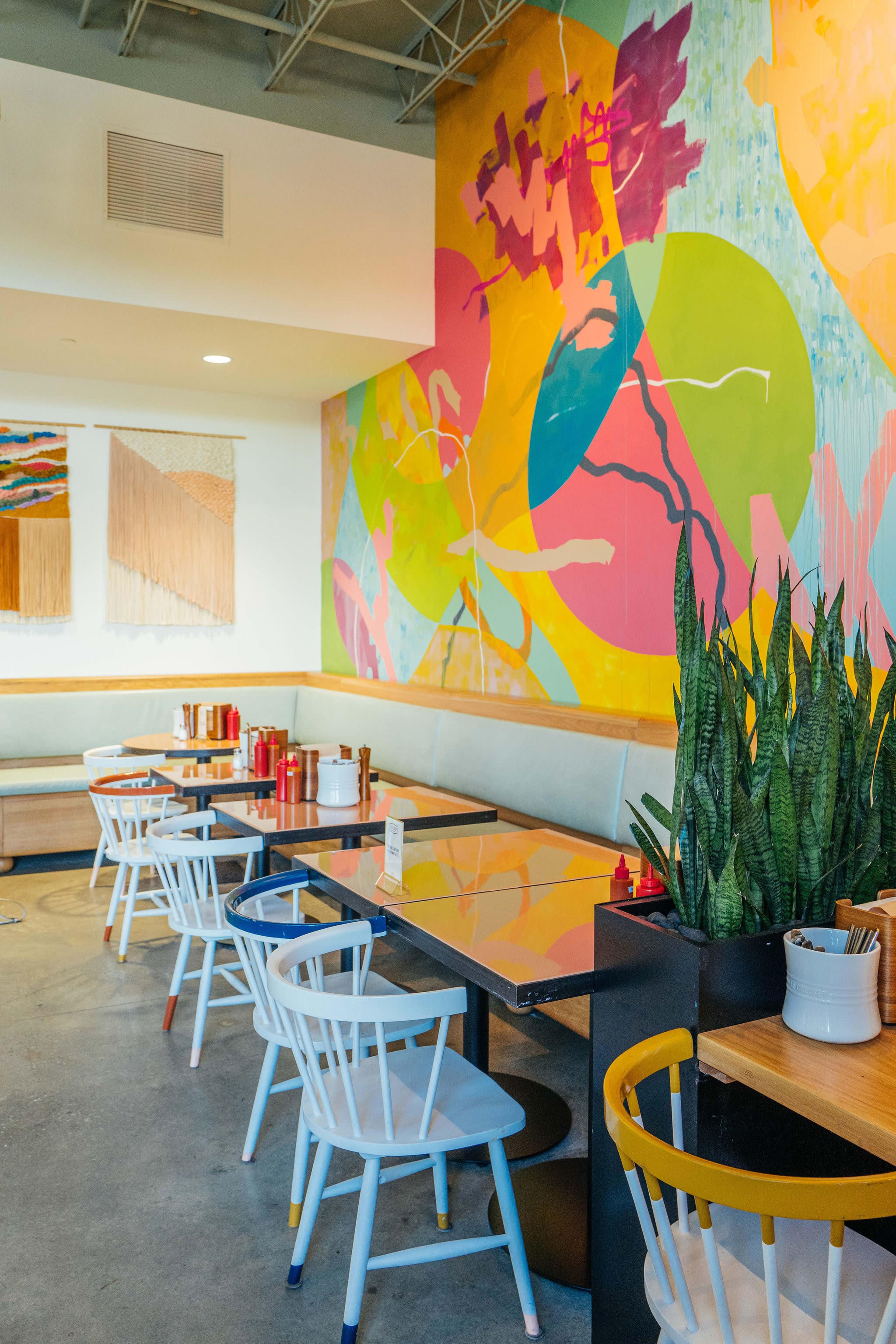 The image shows a modern restaurant interior featuring colorful abstract wall art, wooden tables, and white chairs with a leafy plant in the foreground.