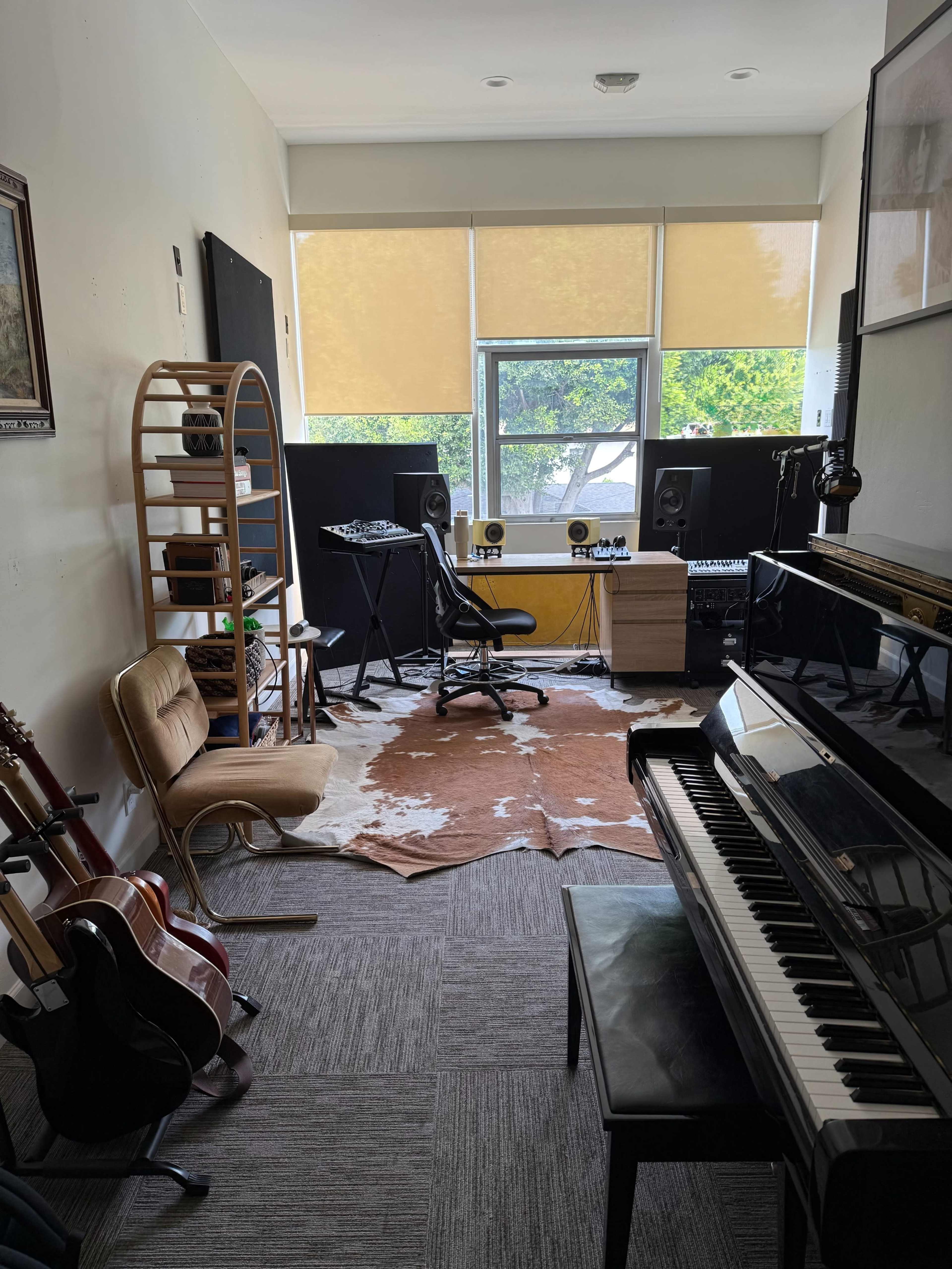 The image shows a small music studio with a piano, guitars, a desk with audio equipment, and large windows letting in natural light.