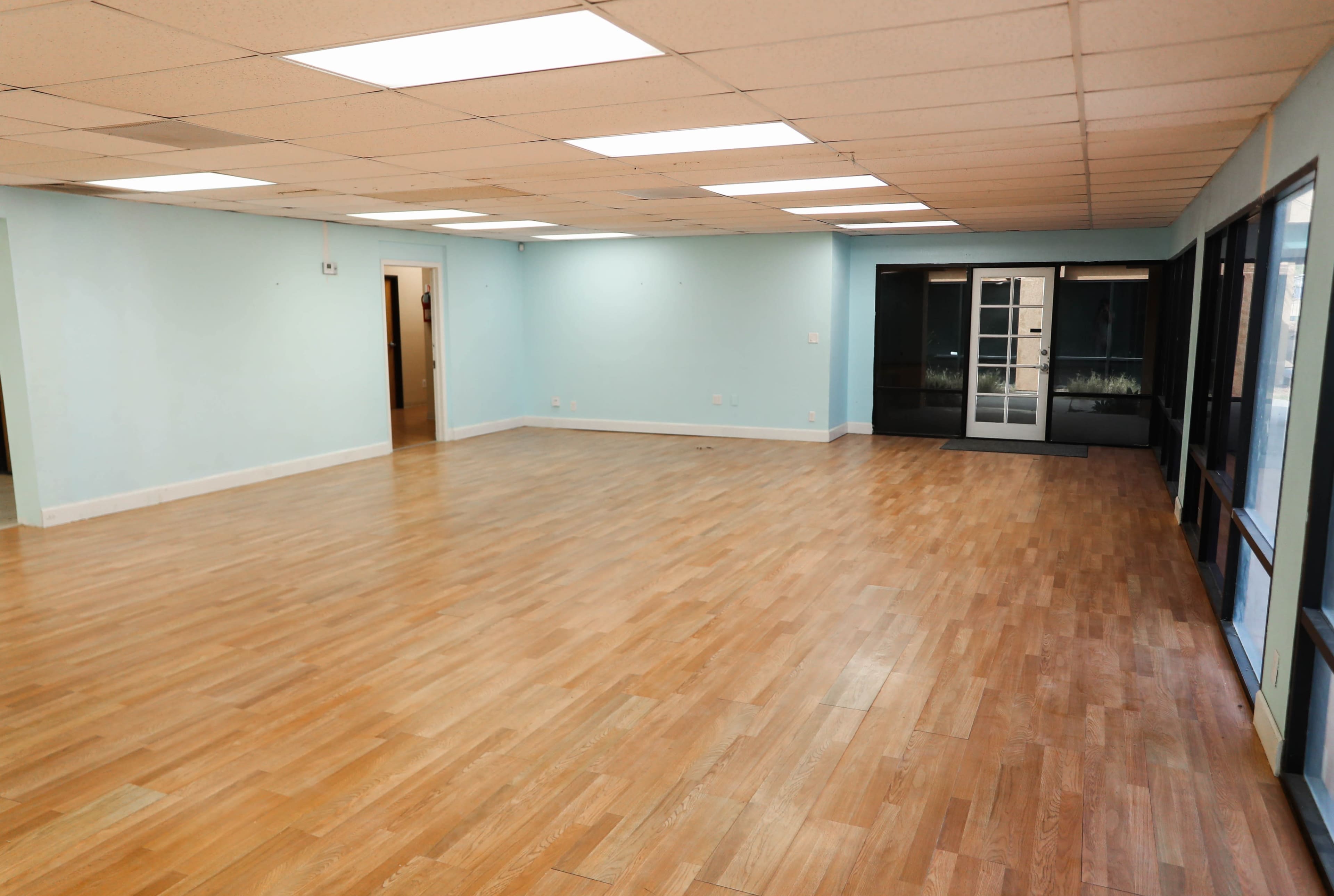 An empty room with light blue walls and hardwood flooring features overhead fluorescent lights and large windows on one side.