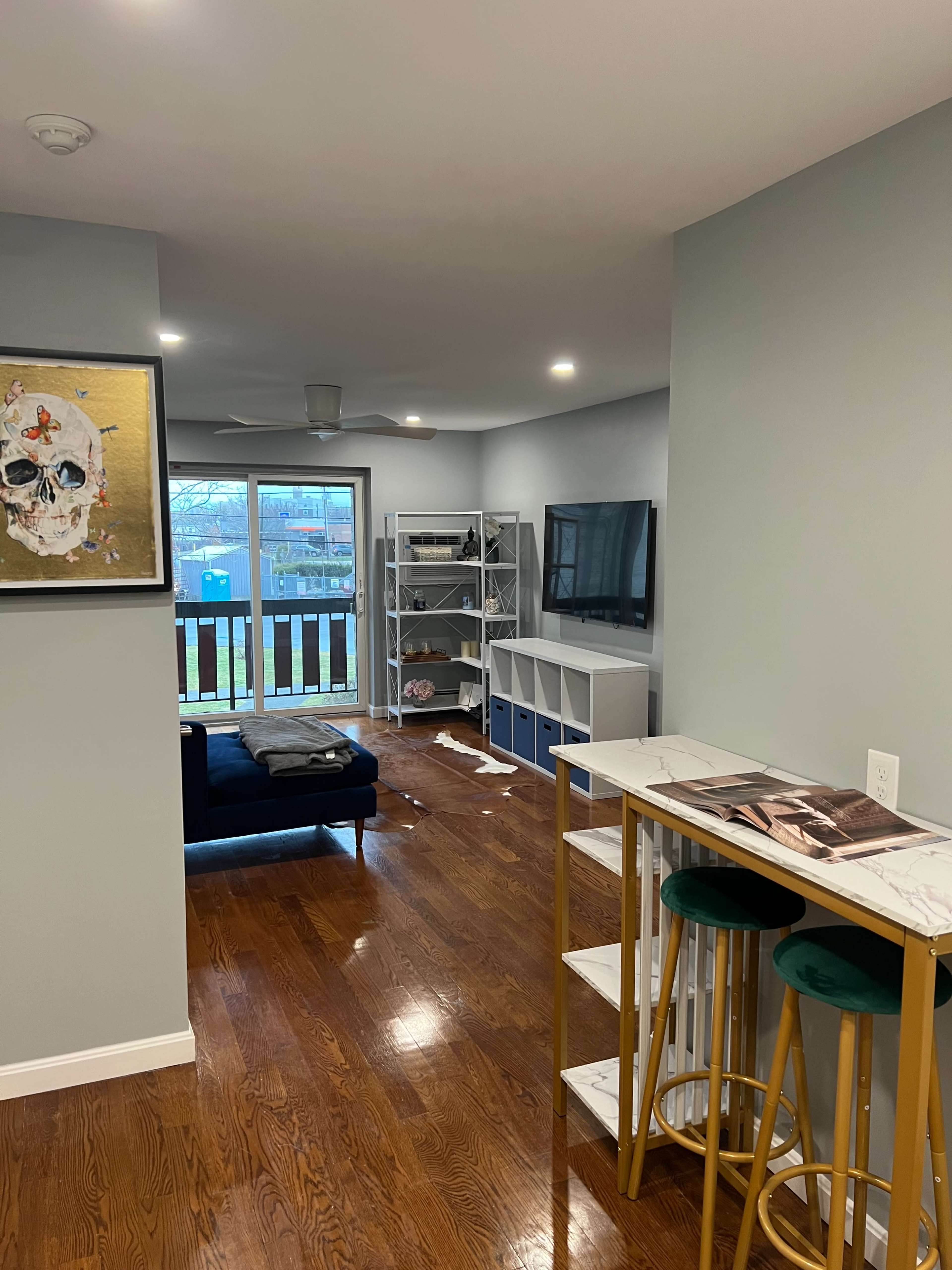 A modern living room with wooden floors, a dark couch, a wall-mounted television, and a small dining area with bar stools.