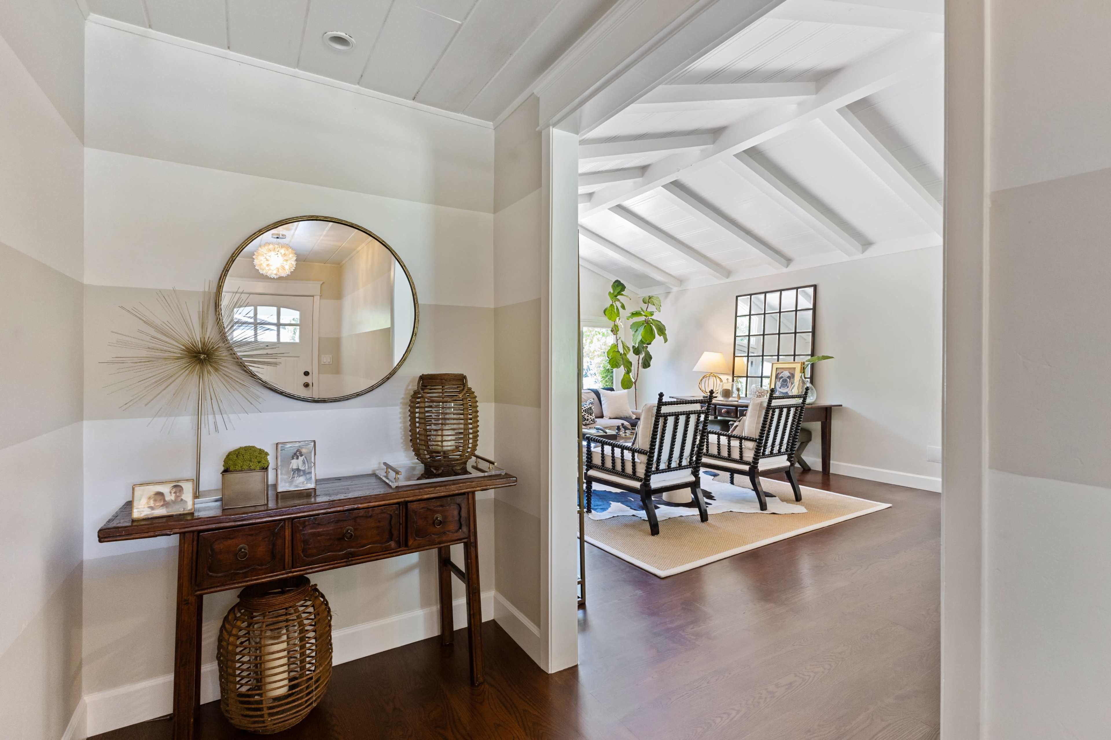 The image shows an interior entryway leading to a living room, featuring a console table with decorative items and a round mirror on the wall.