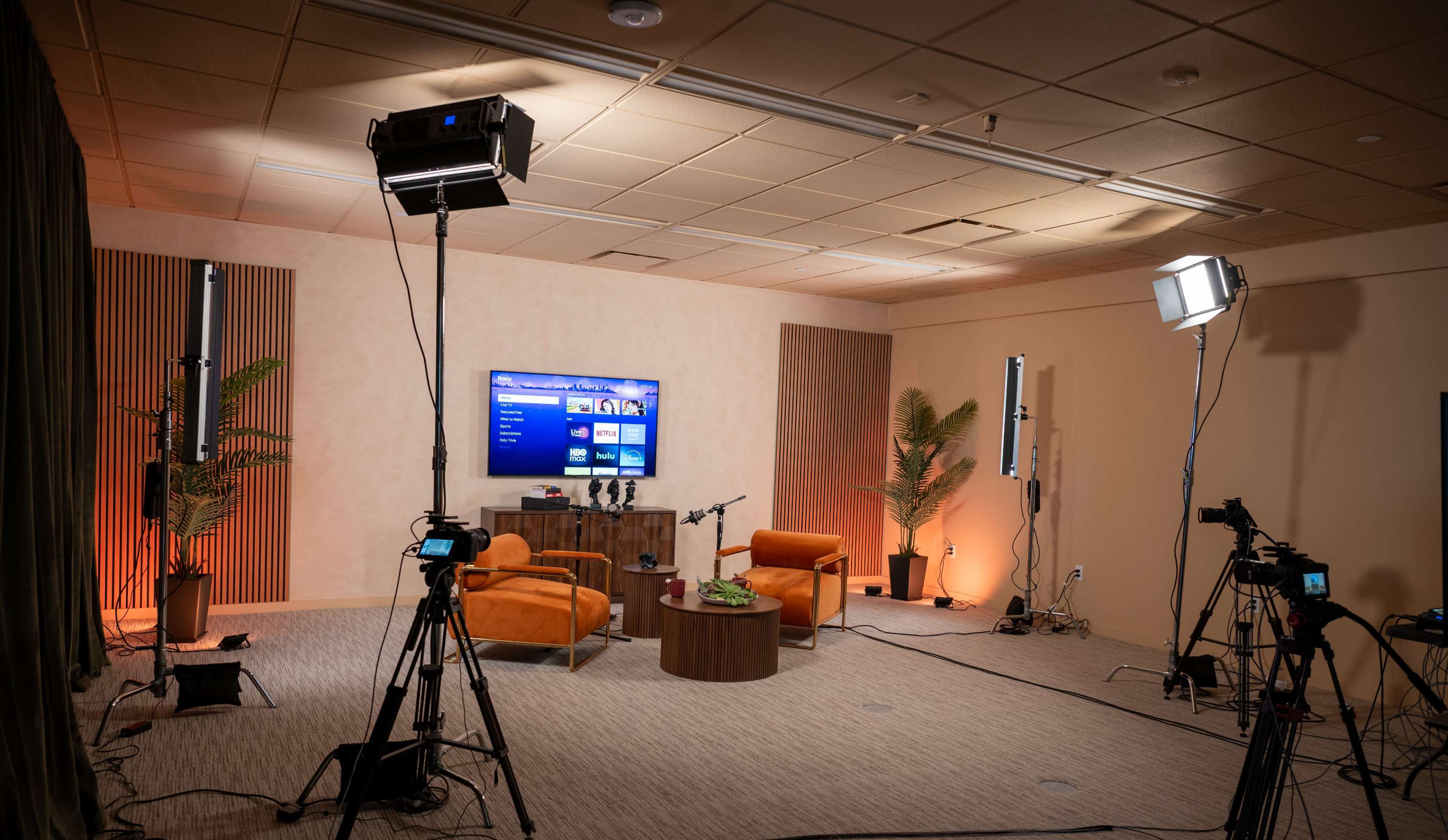 The image shows a well-lit studio setting with two orange chairs, a round table, video cameras, and a large screen displaying content, surrounded by soft lighting and plants.