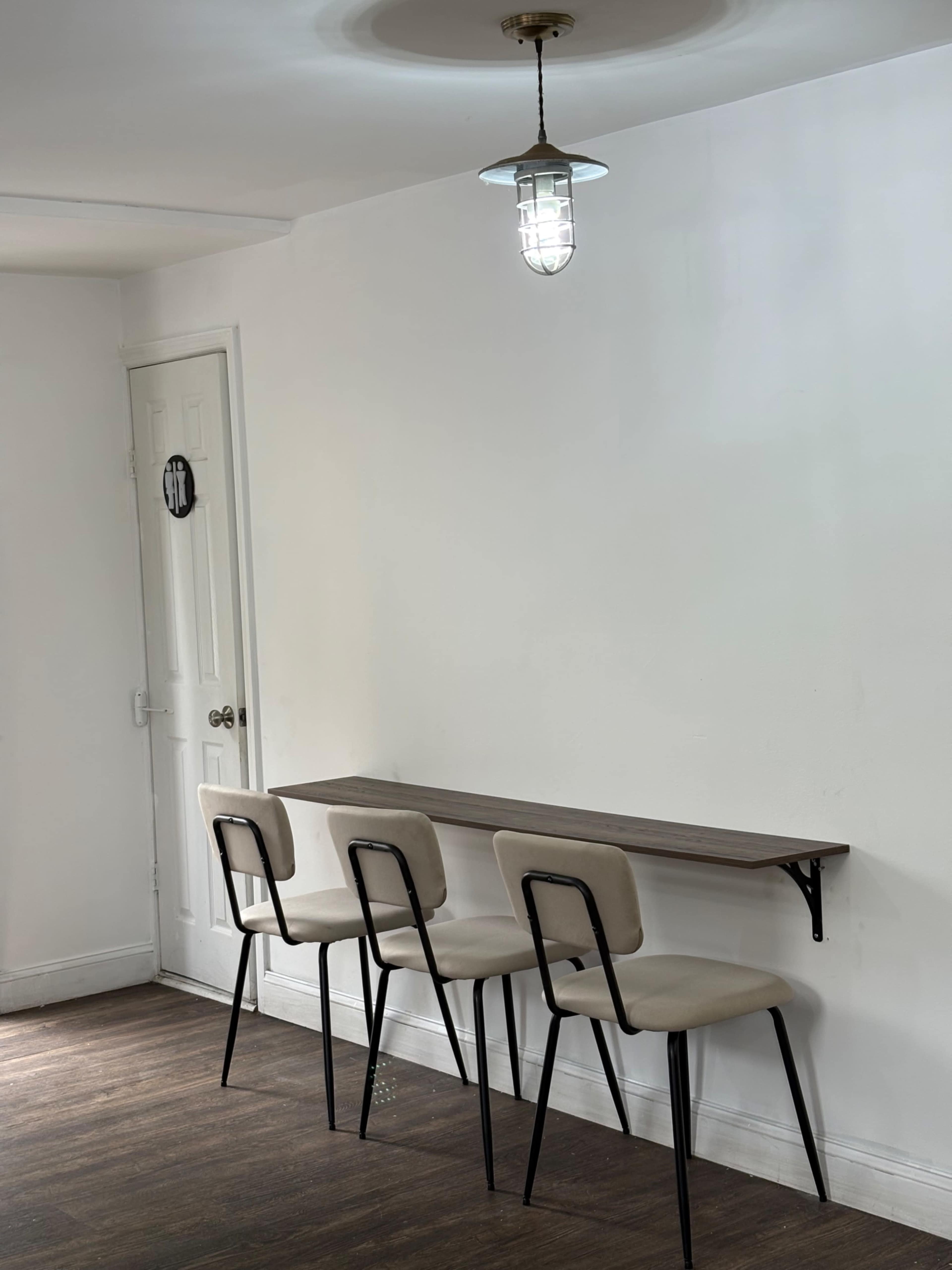 A narrow table with three beige chairs is mounted against a white wall, illuminated by a pendant light above.