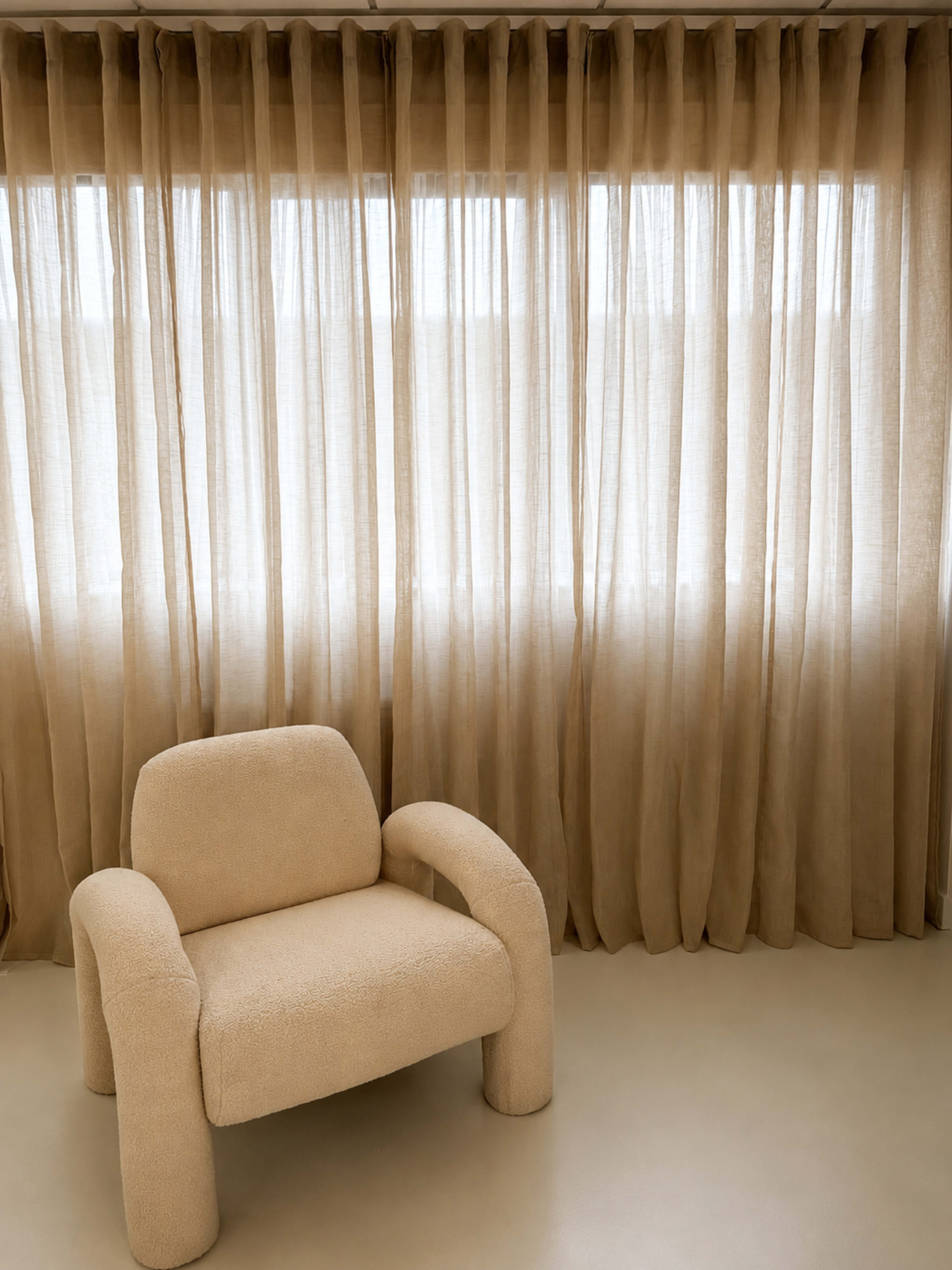A beige, upholstered chair sits in front of sheer, tan curtains near a window.