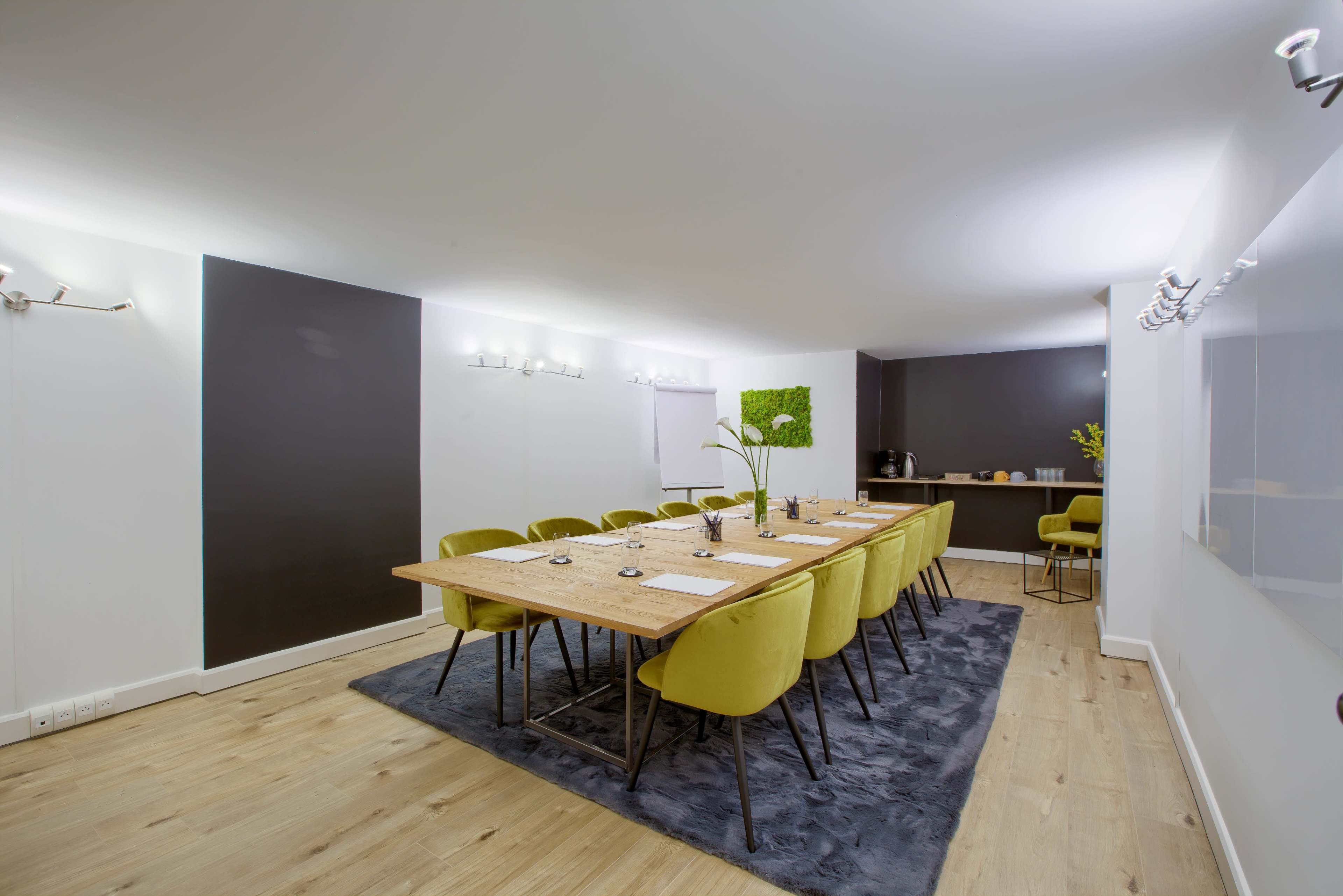 A modern conference room features a long wooden table surrounded by green chairs, with a minimalistic design and soft lighting.